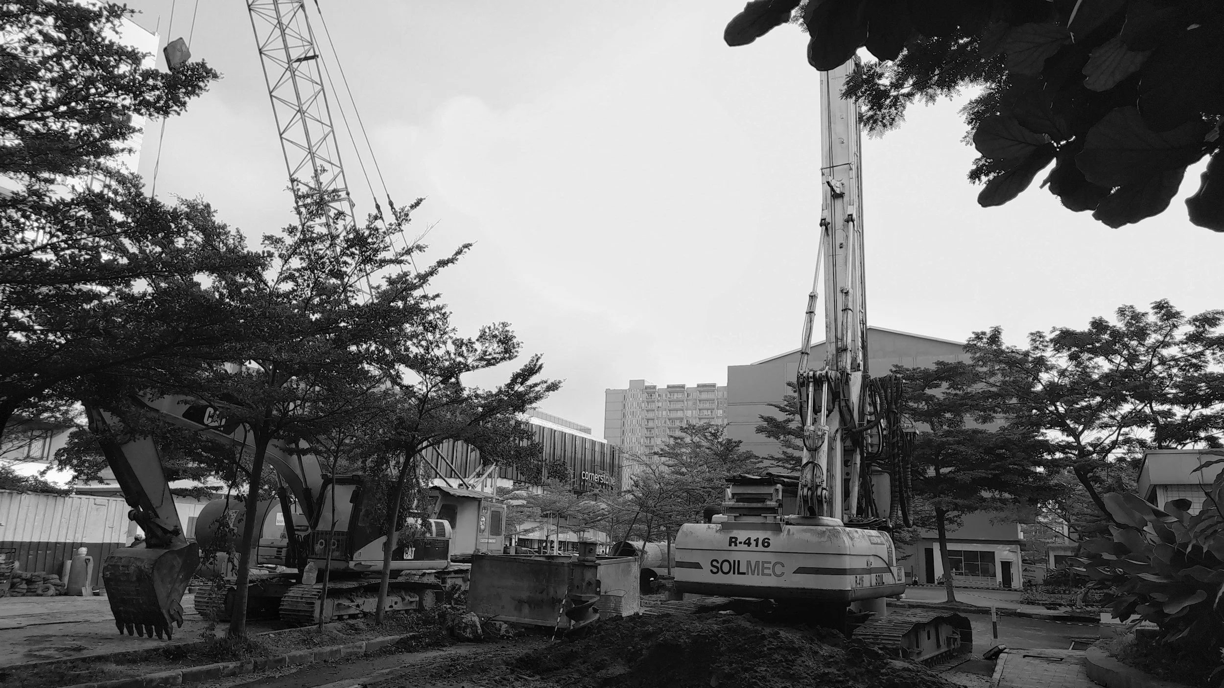 Construction site with excavator and crane in an urban area, surrounded by trees and buildings.