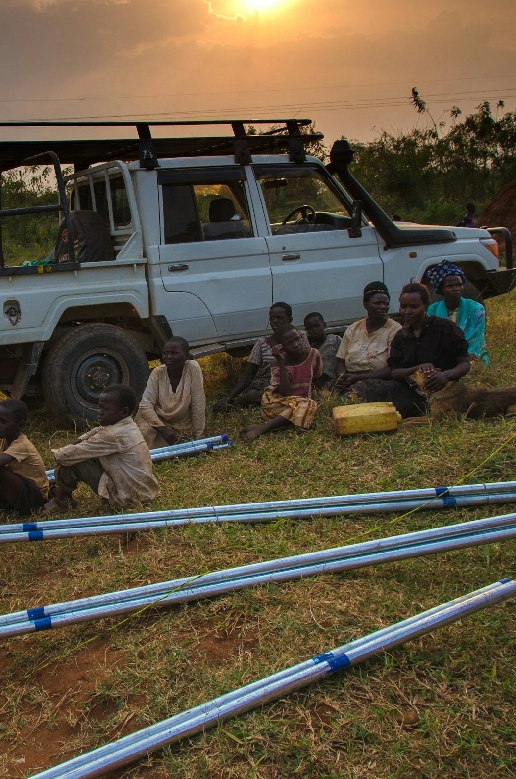 Group of people sitting on grass next to a white pickup truck with a rooftop rack, under a sunset sky, with scattered trees in the background.
