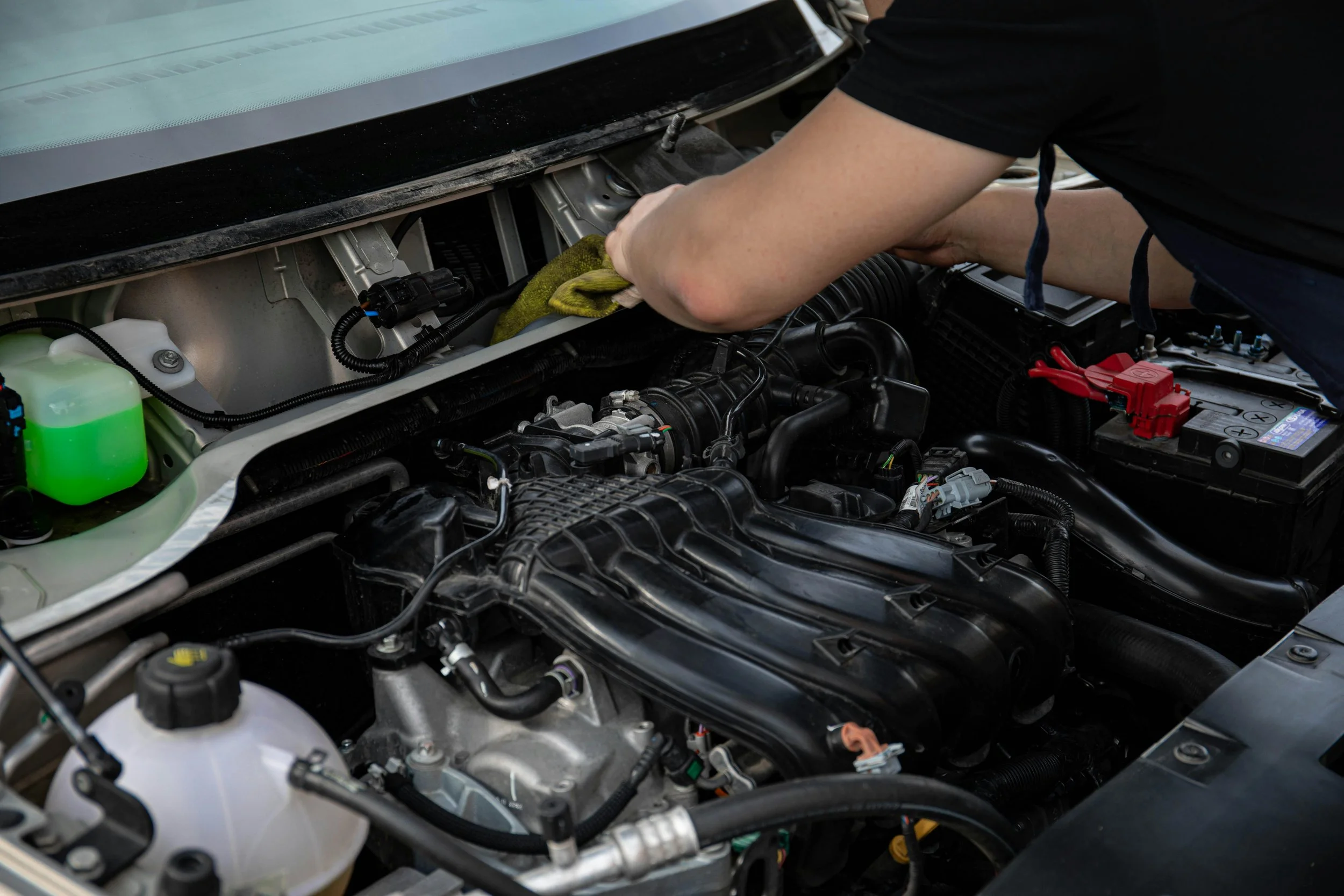 A person working on a car engine, using a yellow cloth, with a visible car battery and various engine components.