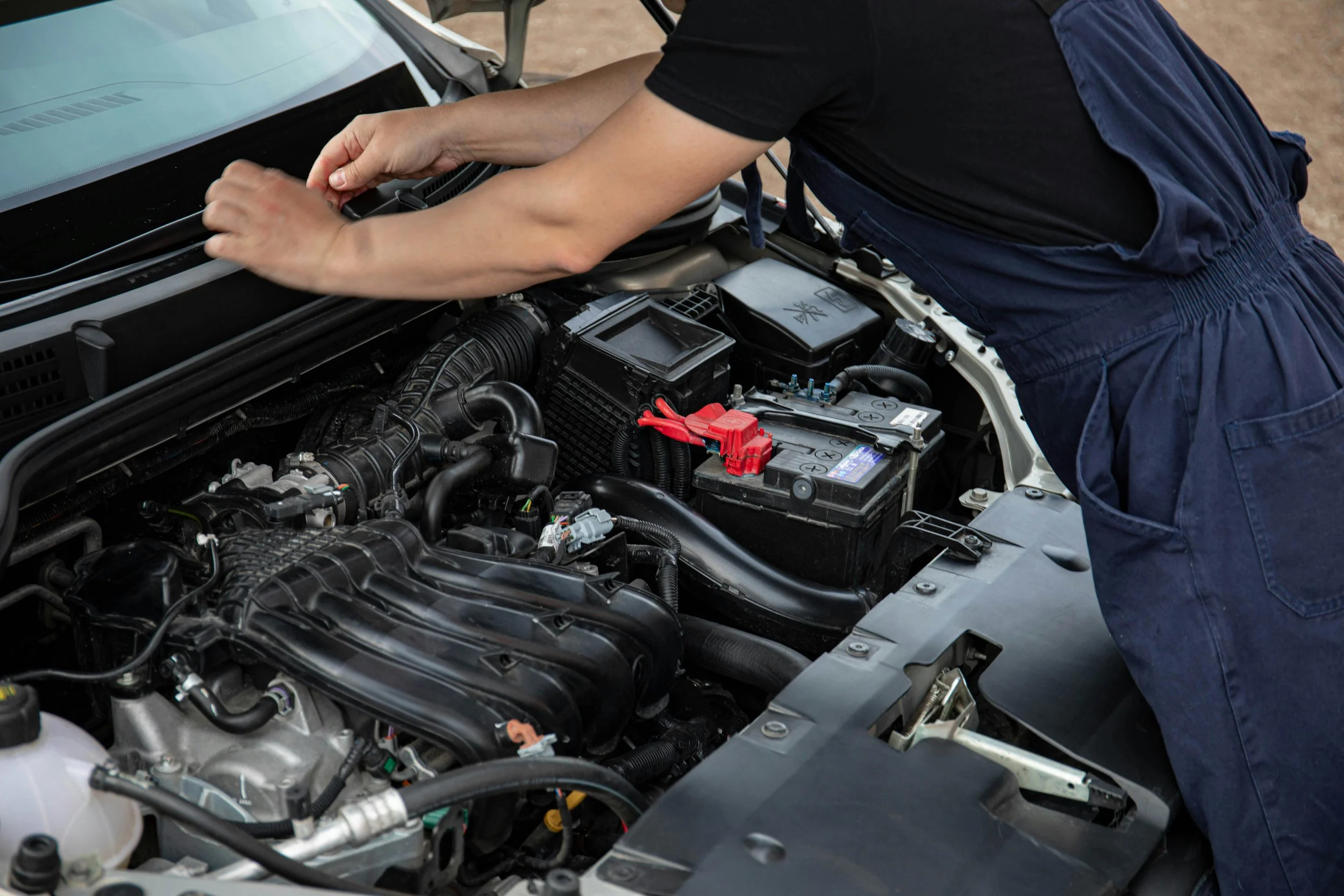 Person working on a car engine in an outdoor setting, wearing a black shirt and dark blue overalls, inspecting components under the hood.