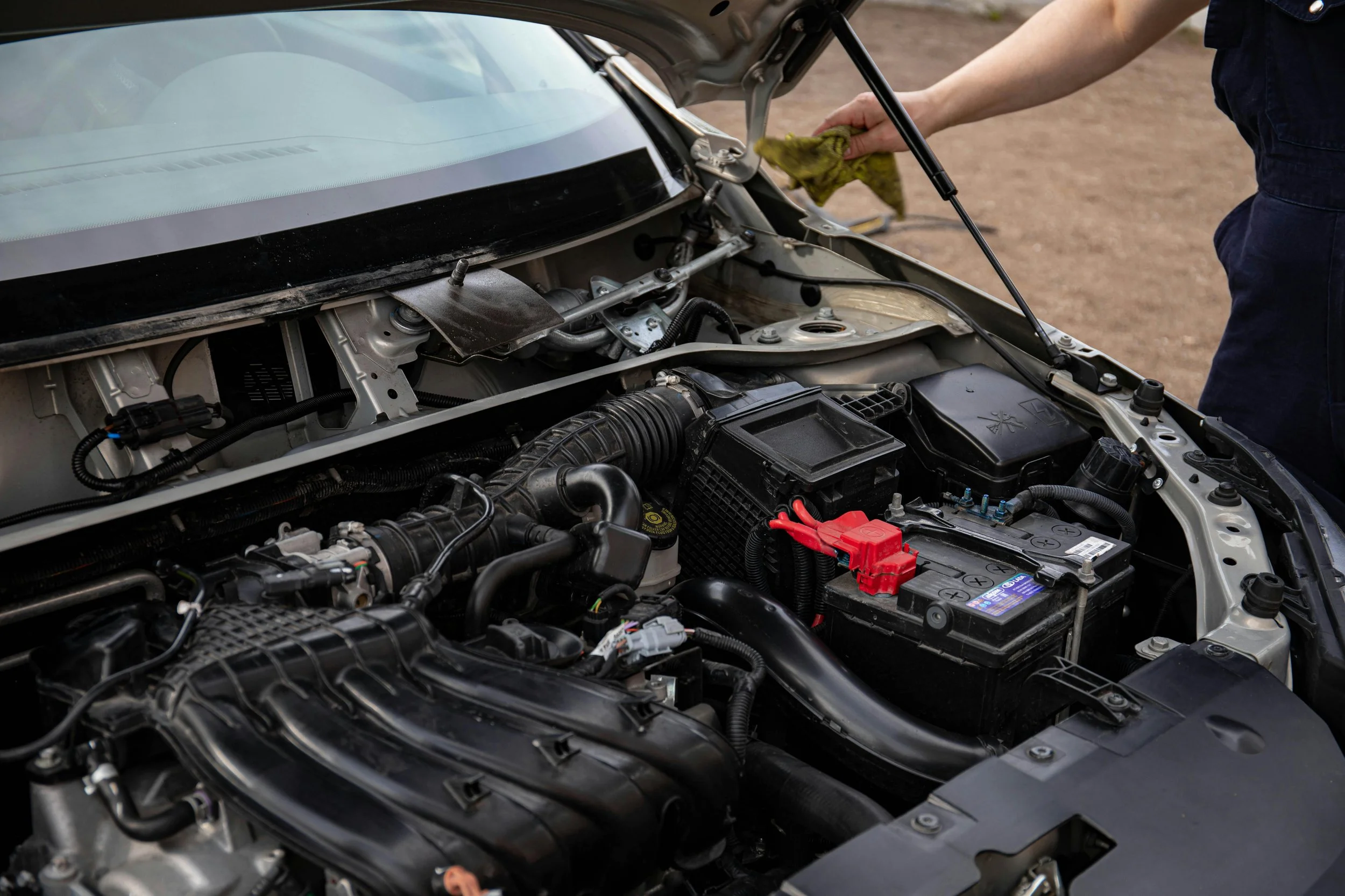 Person inspecting car engine with hood open, holding a cloth in their right hand.