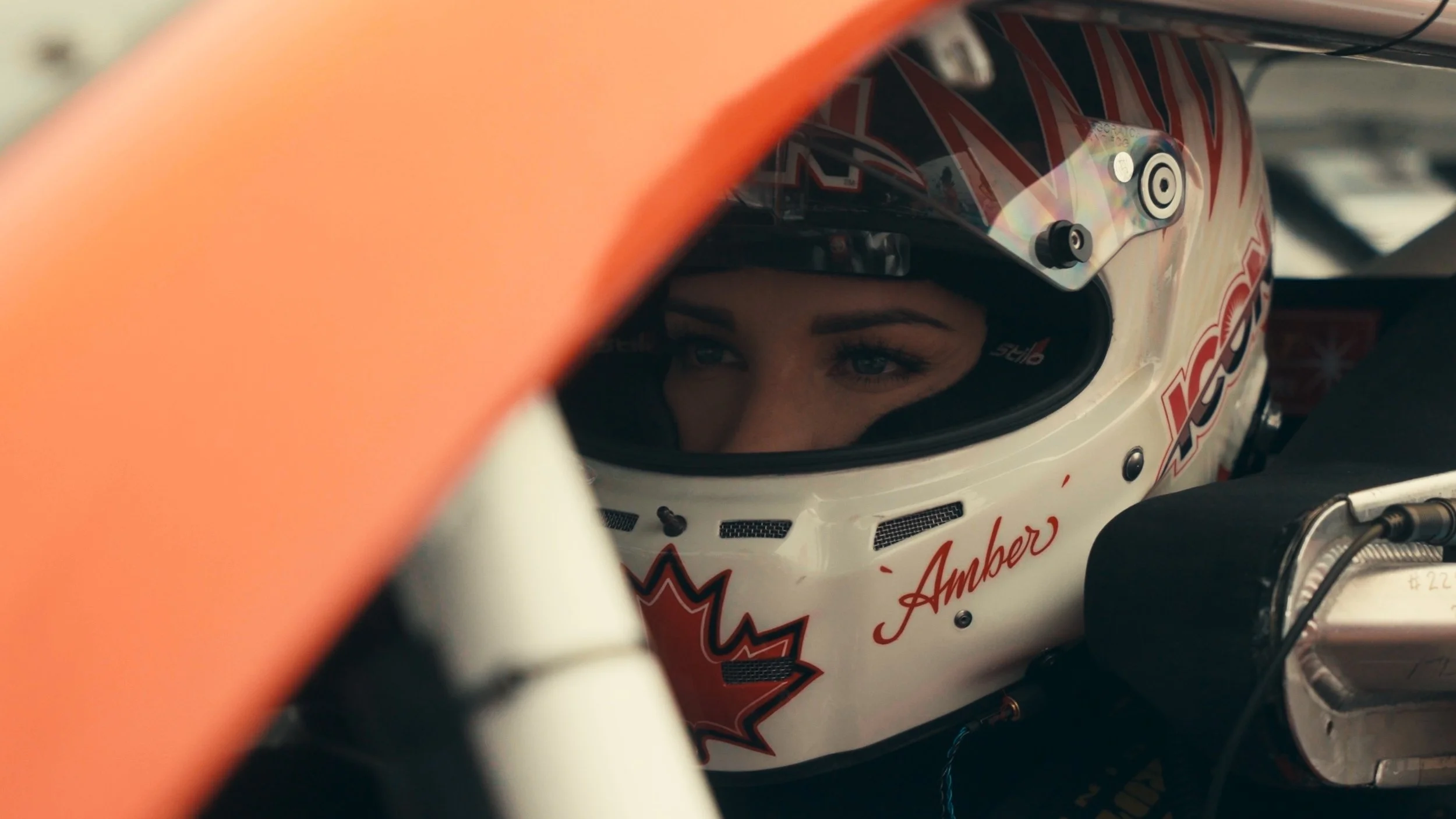 A close-up of a race car driver inside her helmet, looking out through the visor, with part of the car’s body visible in the foreground.