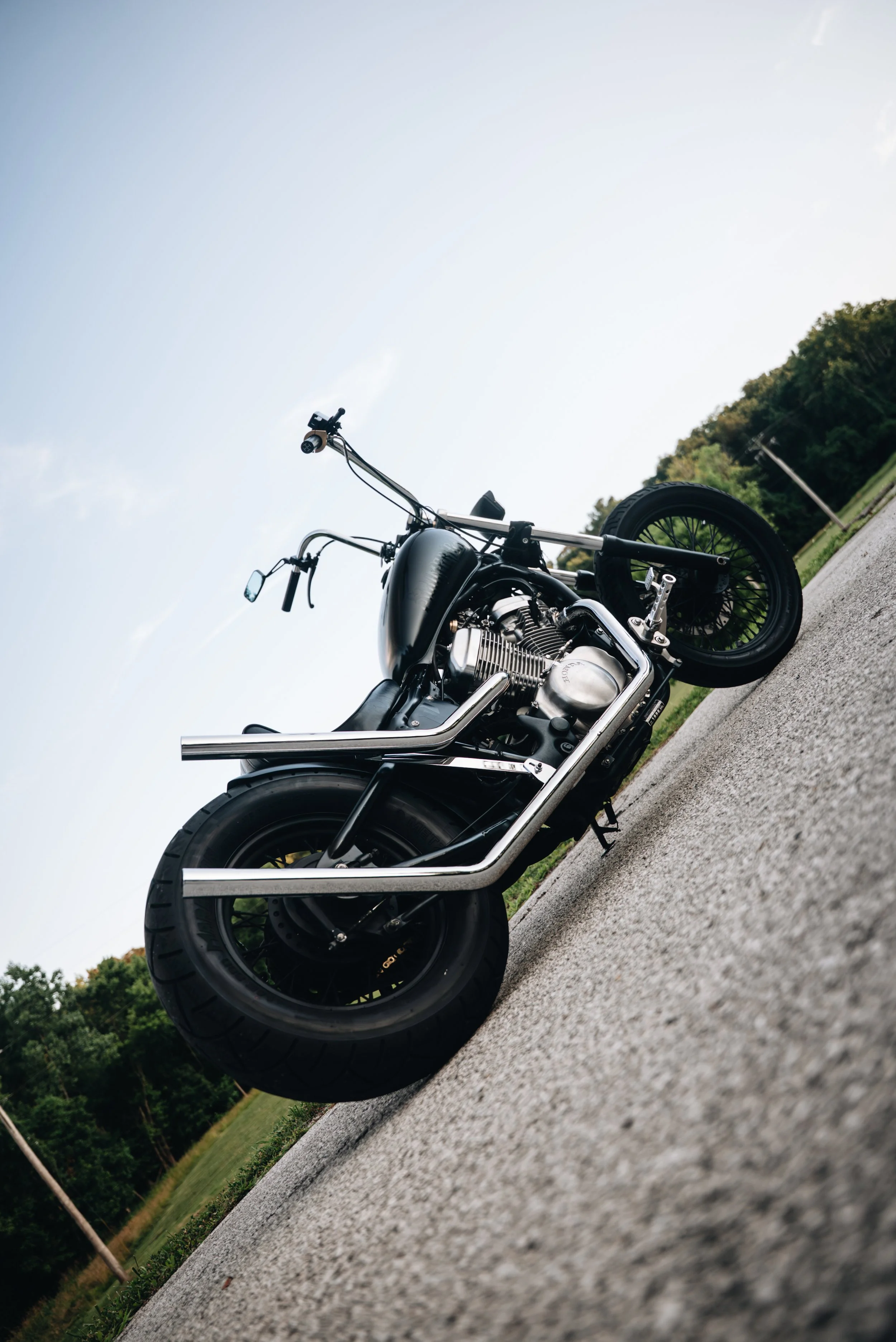 A black motorcycle parked on a paved road, with a background of trees and a grassy field under a clear sky.
