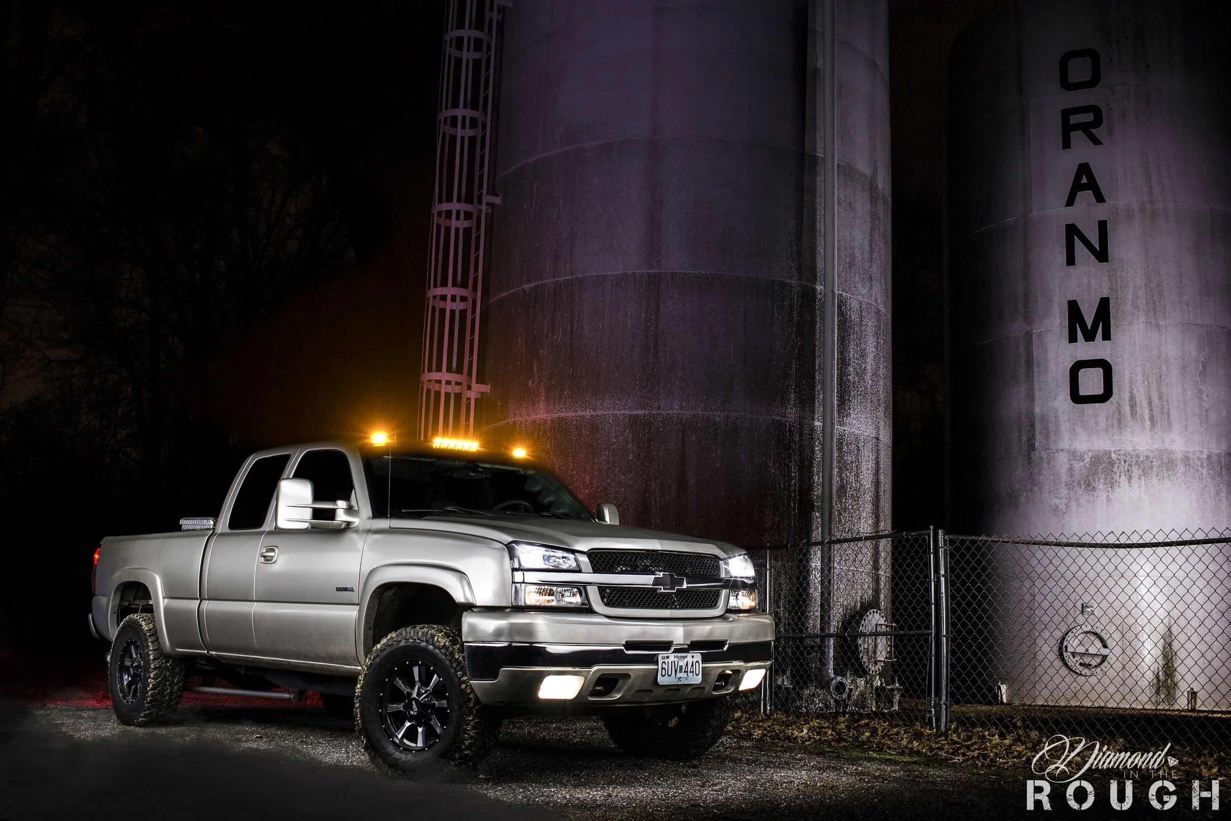 A silver pickup truck with large tires and black rims parked near a large industrial storage tank at night, illuminated by a streetlight and truck lights.
