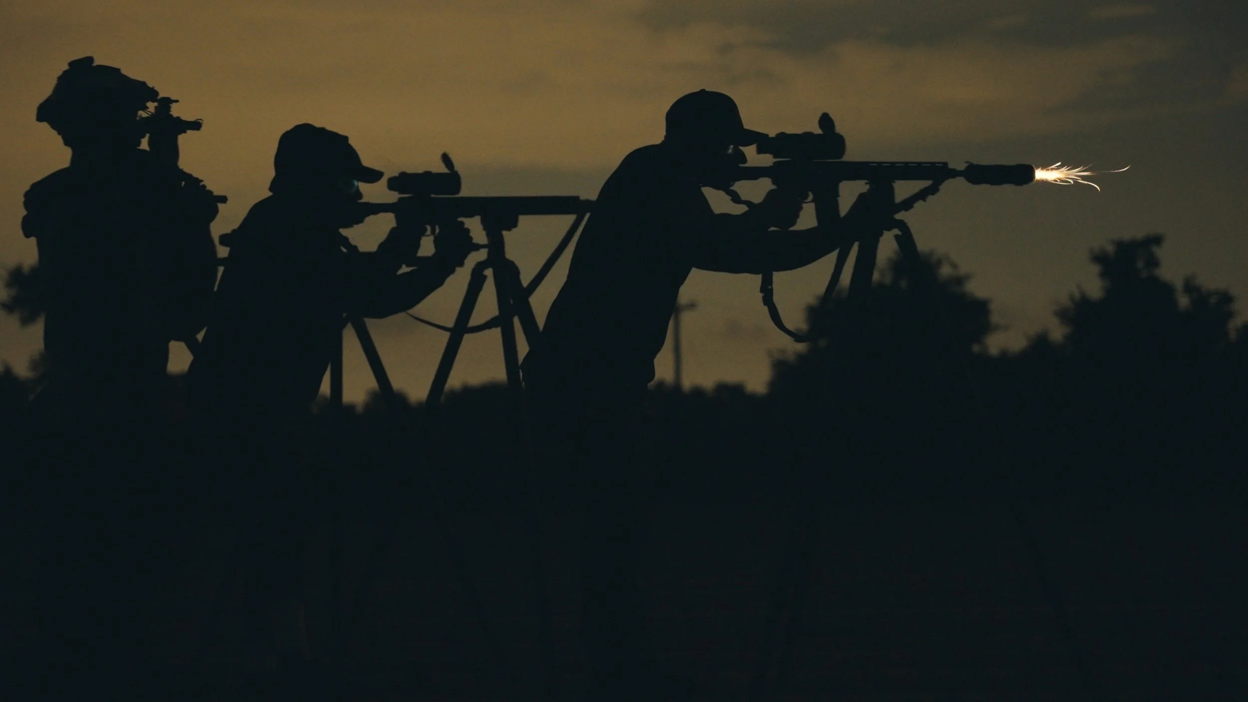 Silhouettes of four soldiers aiming rifles with attached scopes and bipods, firing at night, with a faint glow from the muzzle of the rifle on the right.