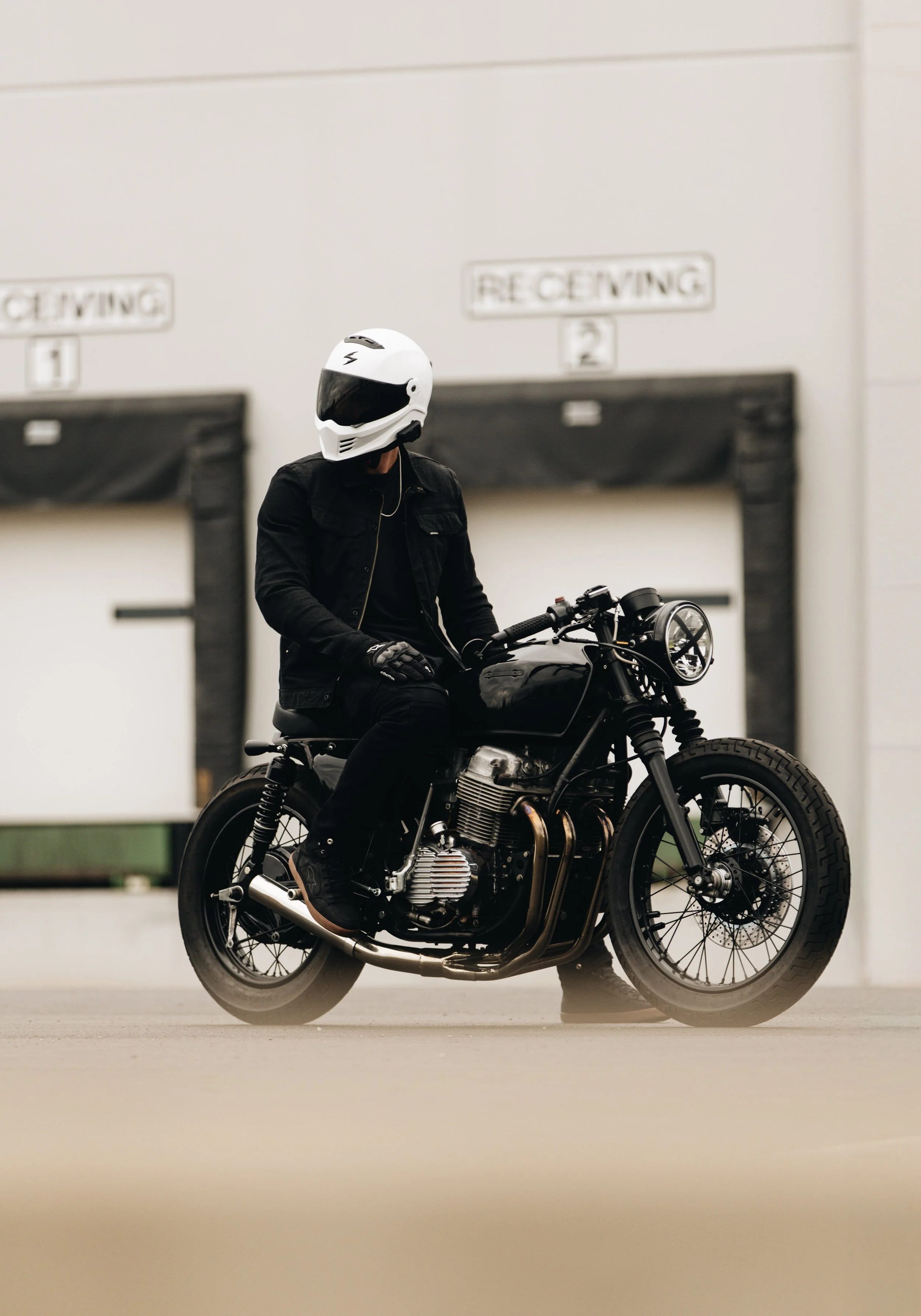 Person wearing a white helmet with a black visor, riding a black vintage style motorcycle in front of a white wall with 'RECEIVING' signs.