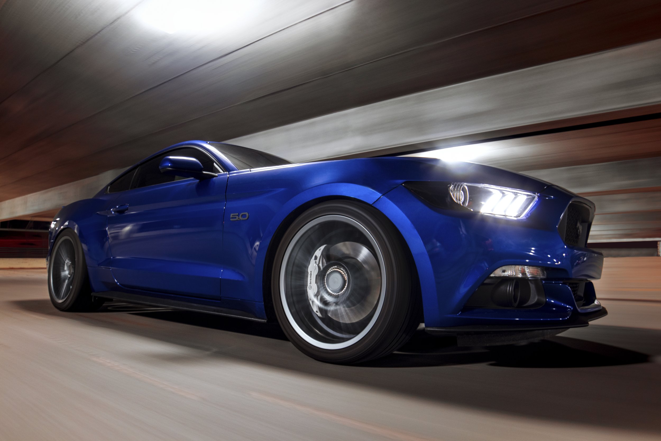 A blue sports car, a Ford Mustang 5.0, driving through a tunnel with a motion blur effect, creating a sense of speed.