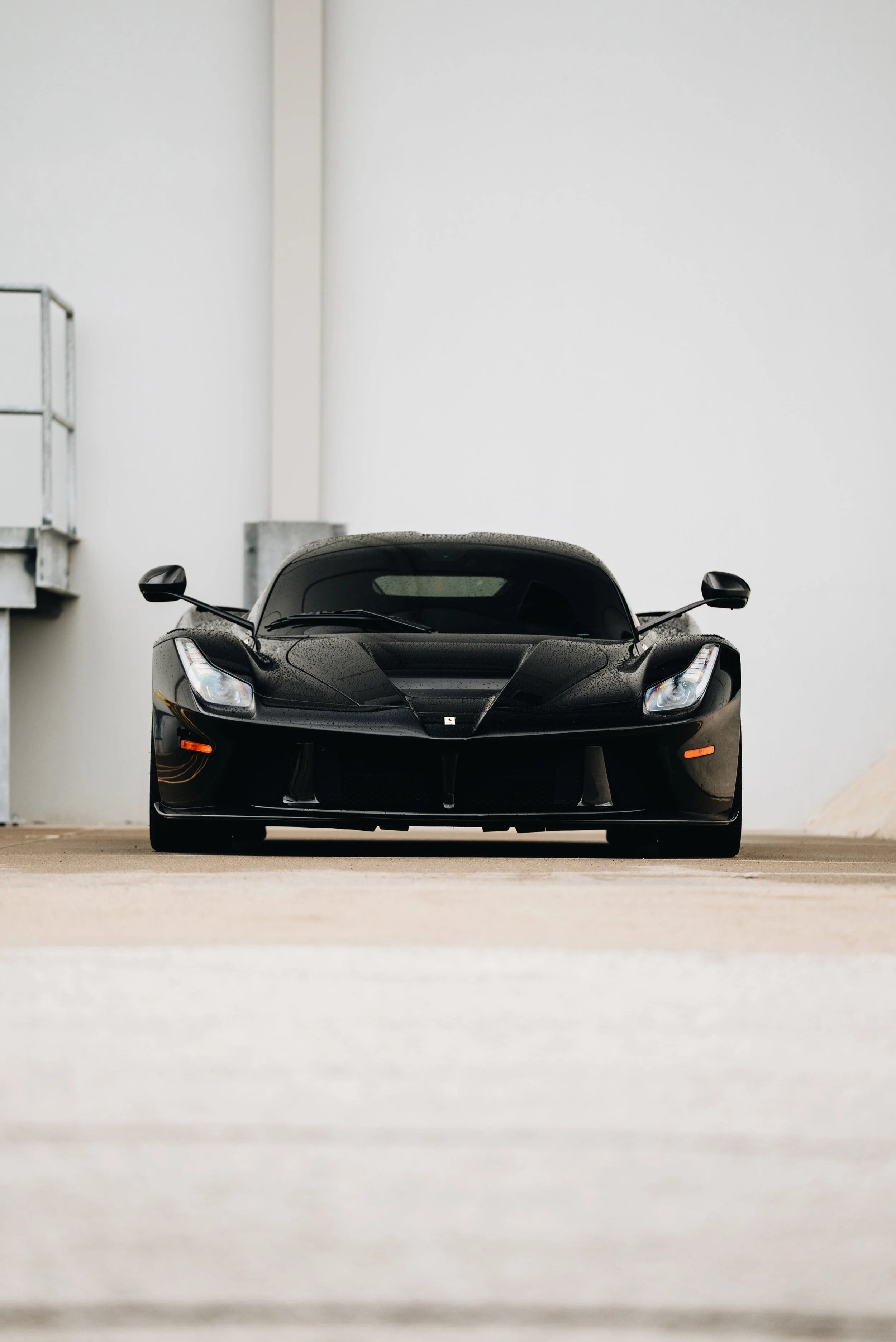 Front view of a black sports car parked in a minimalistic space with a white wall in the background.