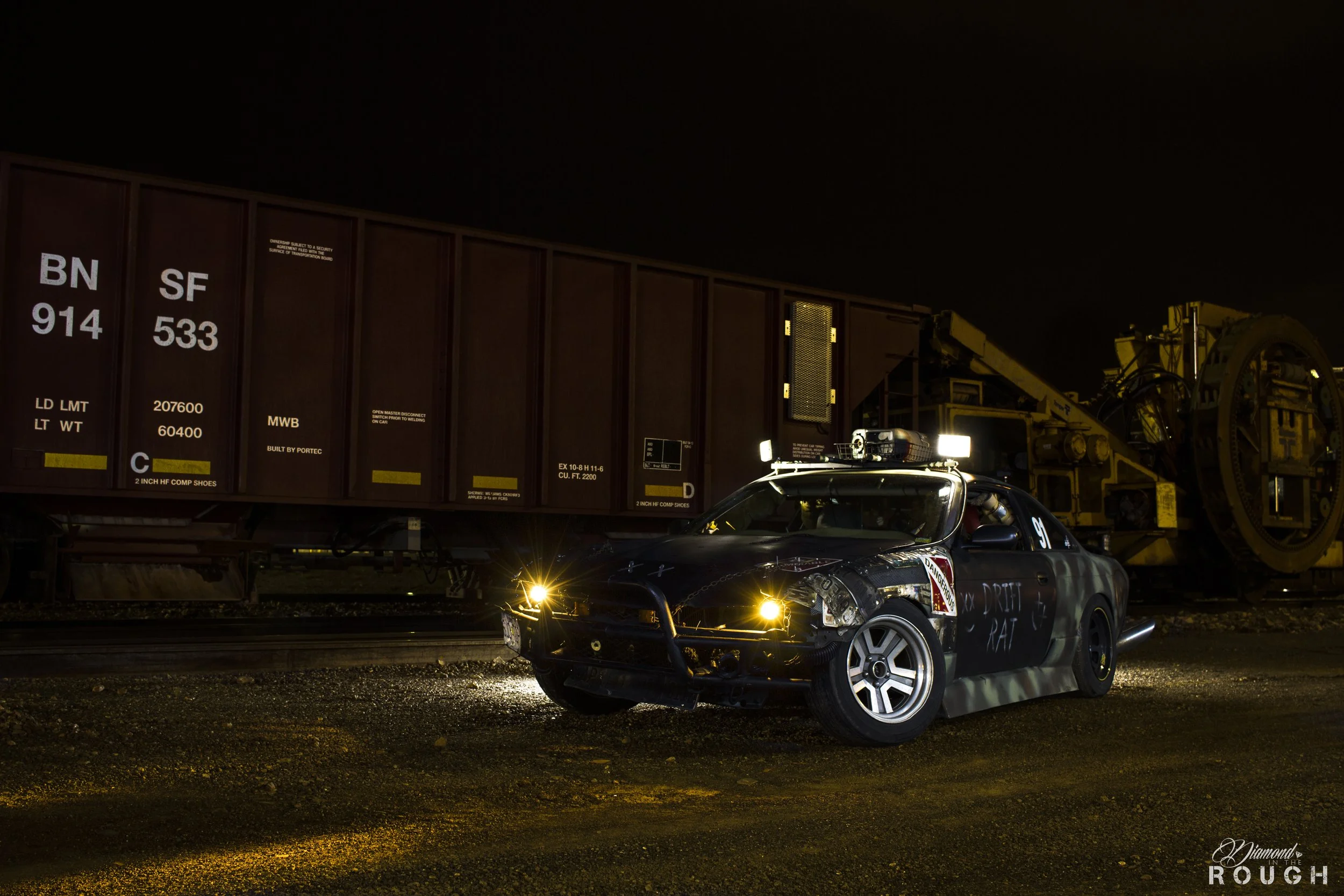 Night scene of a damaged car with part of the front bumper missing and graffiti on the side, parked next to a train car on a railway track. Bright lights illuminate the vehicles, with a dark sky in the background.