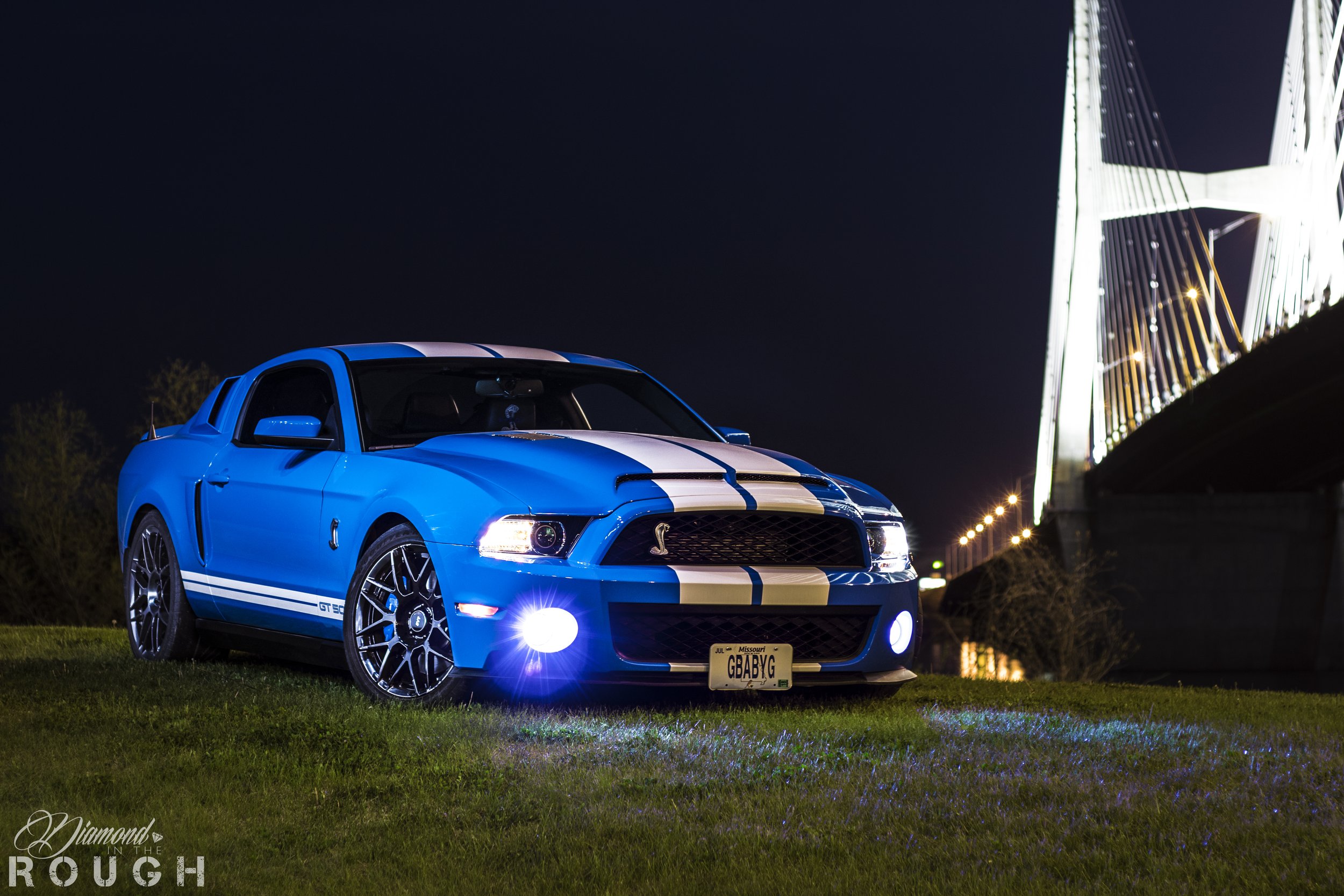 A blue Ford Mustang Shelby GT500 with white racing stripes parked on grass at night, with a illuminated bridge in the background.