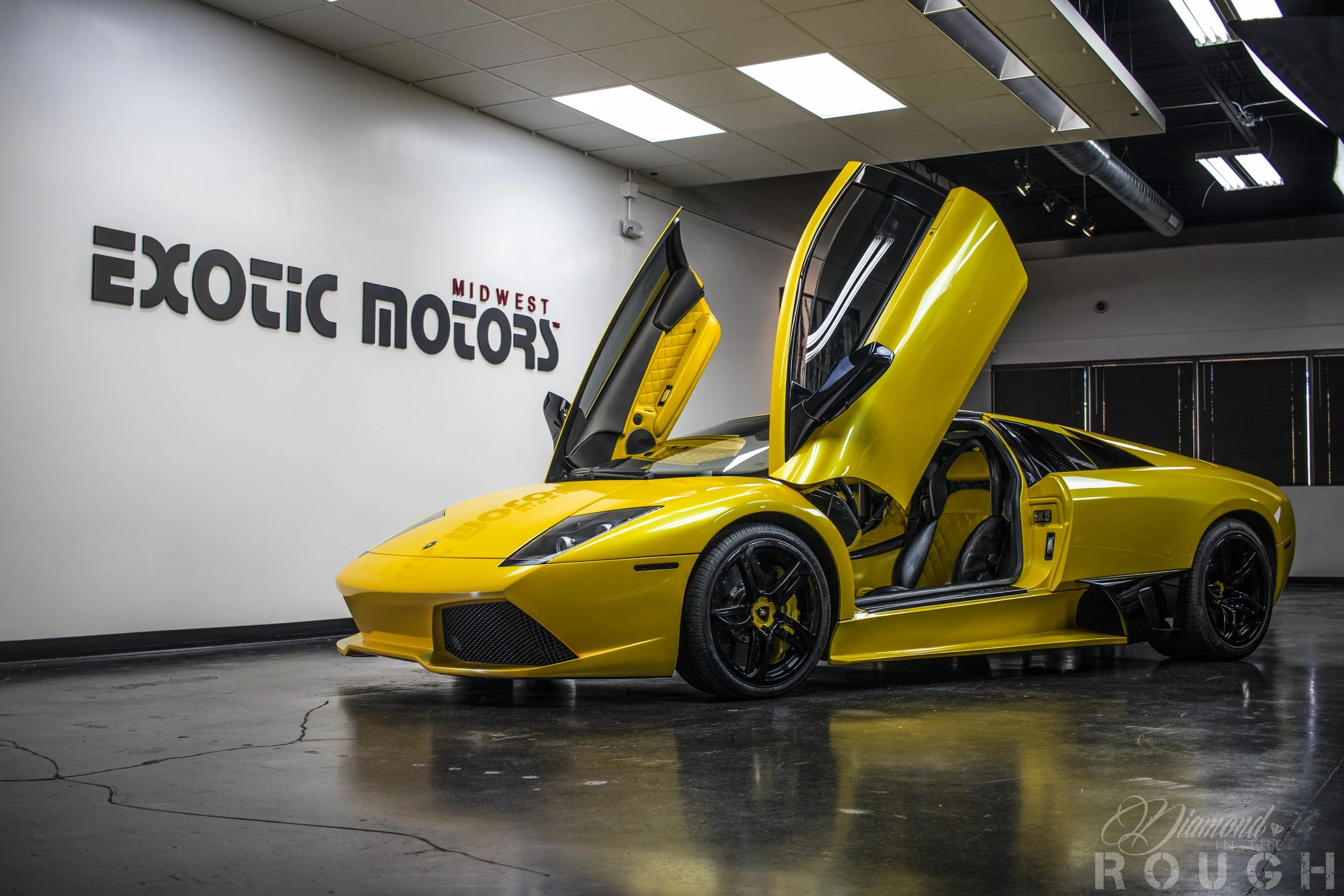 A bright yellow sports car with gull-wing doors open, parked indoors against a white wall with the words 'Exotic Motors Midwest' on it.