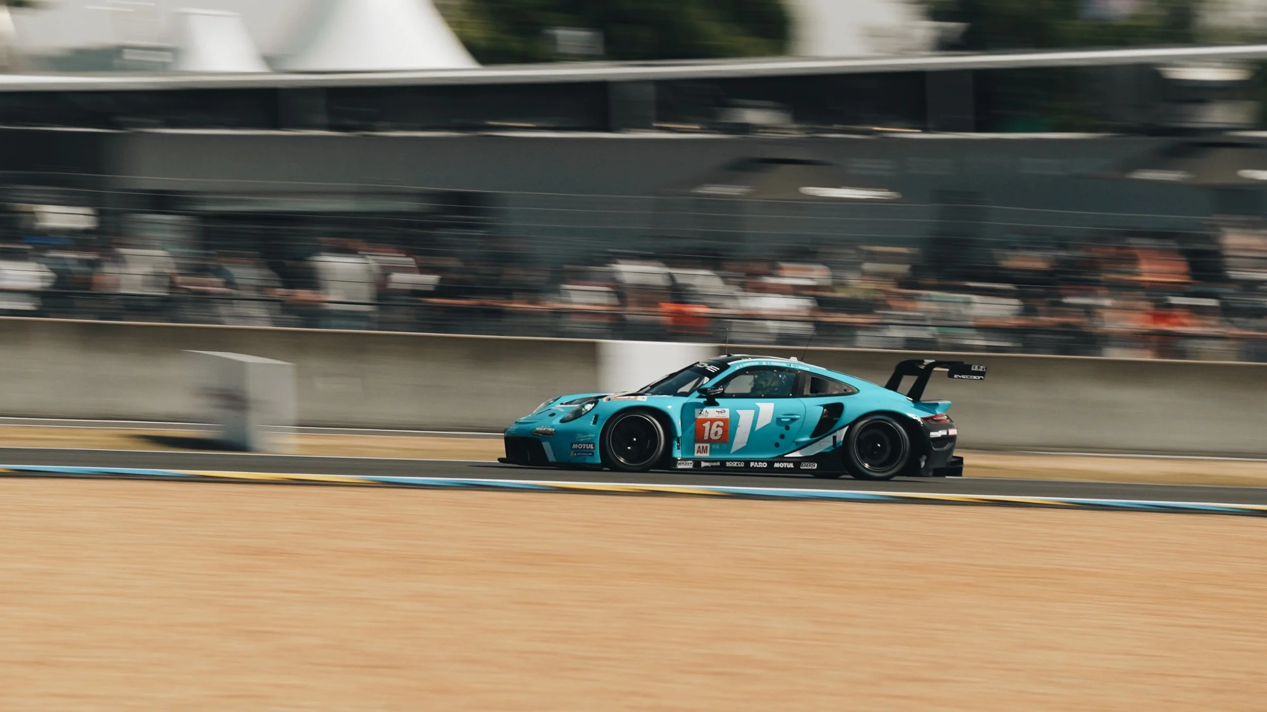 A blue racing car speeding on a race track with a blurred crowd and grandstand in the background.
