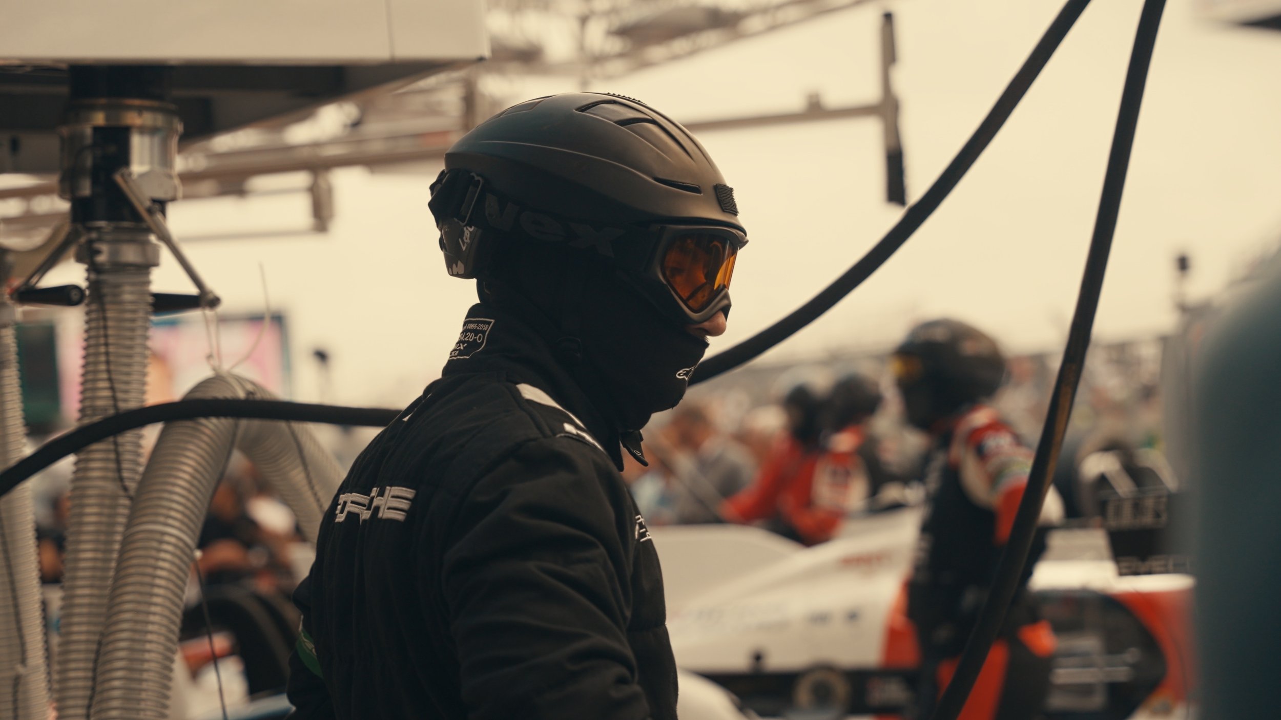 A race car driver wearing a black helmet with orange goggles and black racing suit, standing in a pit garage with racing team members blurred in the background.