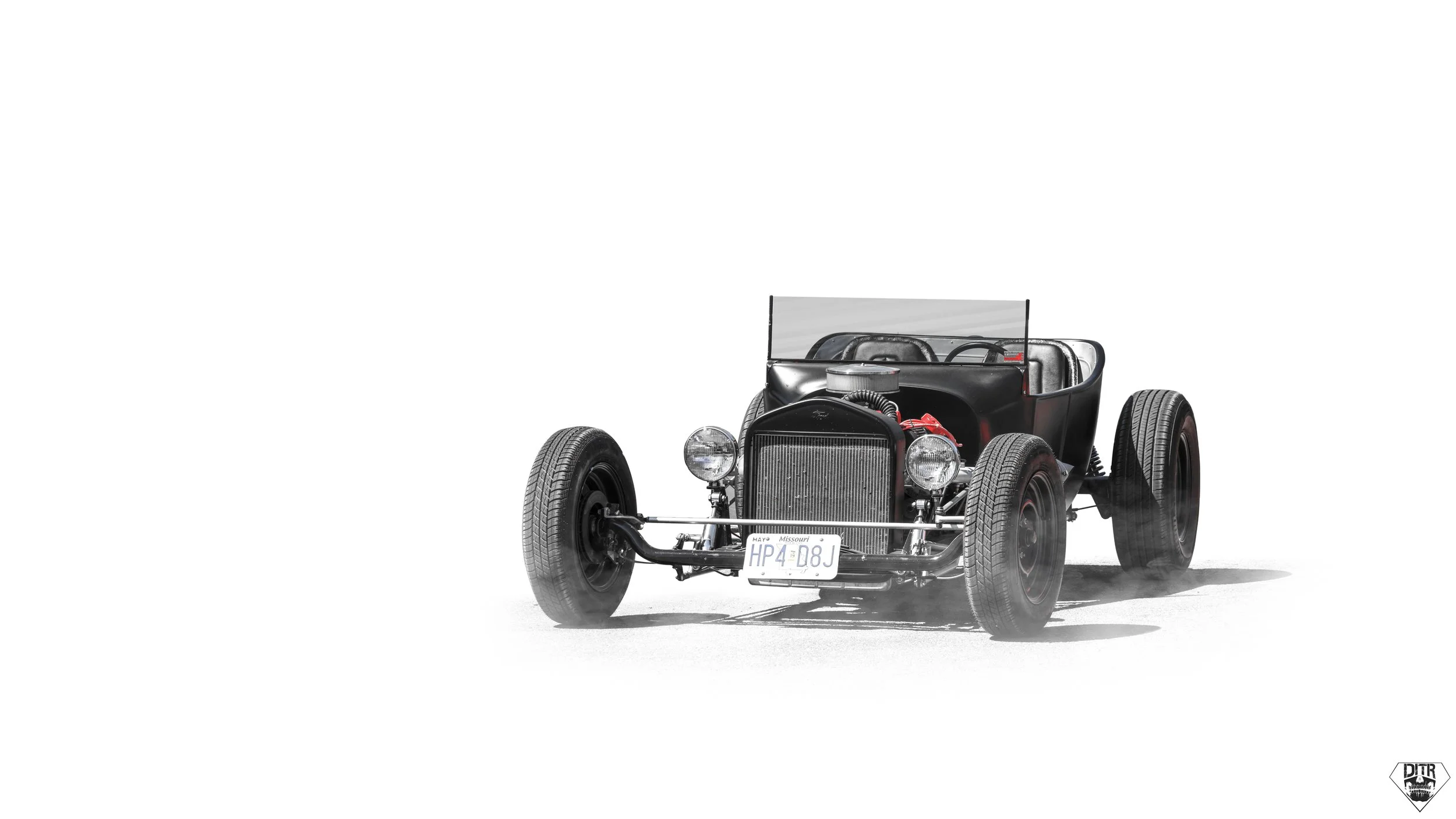 Black vintage race car with a Missouri license plate HP4 D8J, wire-spoked wheels, exposed engine components, and a windshield, on a white background.