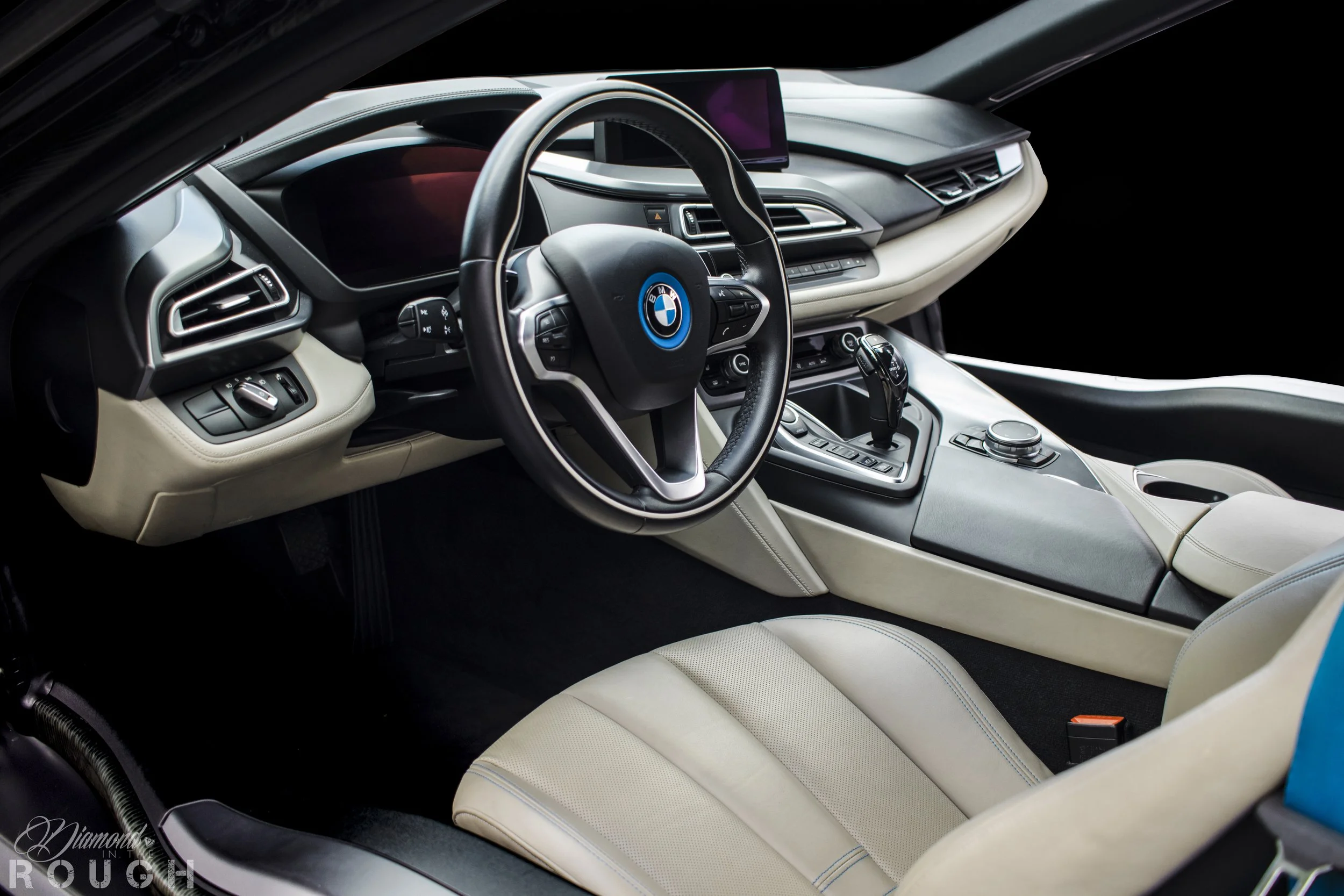 Interior of a BMW car showing the steering wheel, dashboard, and center console with gear shifter and multimedia controls, featuring a modern design with black and beige tones.