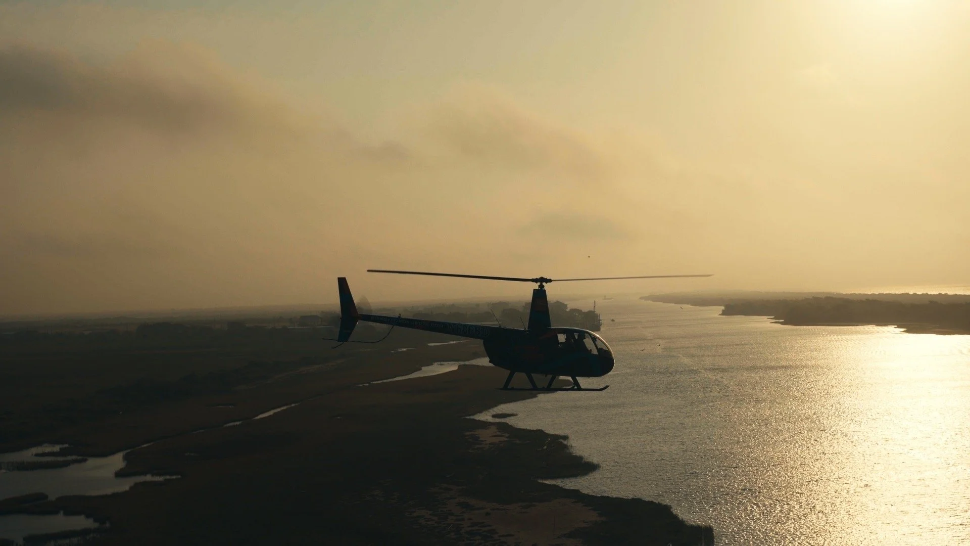 A helicopter parked on a launch pad near a large body of water with a distant shoreline, during sunset or sunrise, with a cloudy sky and golden lighting.