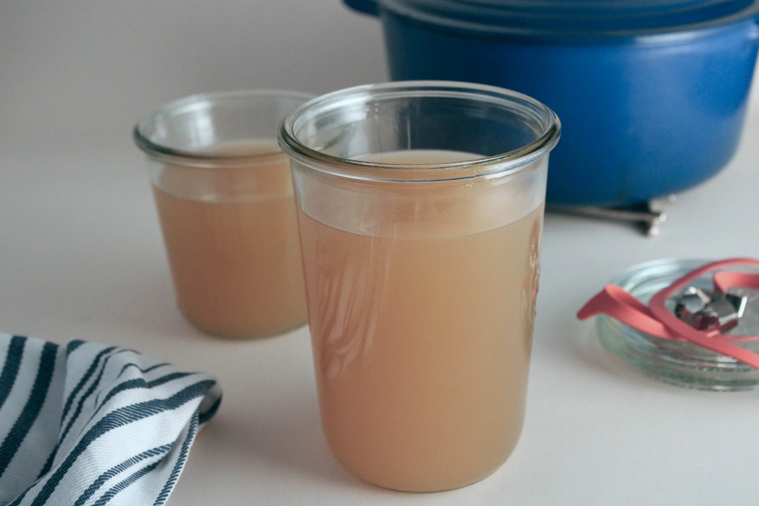 Two glasses of pink beverage, a blue container in the background, part of a striped cloth, and a small dish with pink paper and silver clips on a white surface.