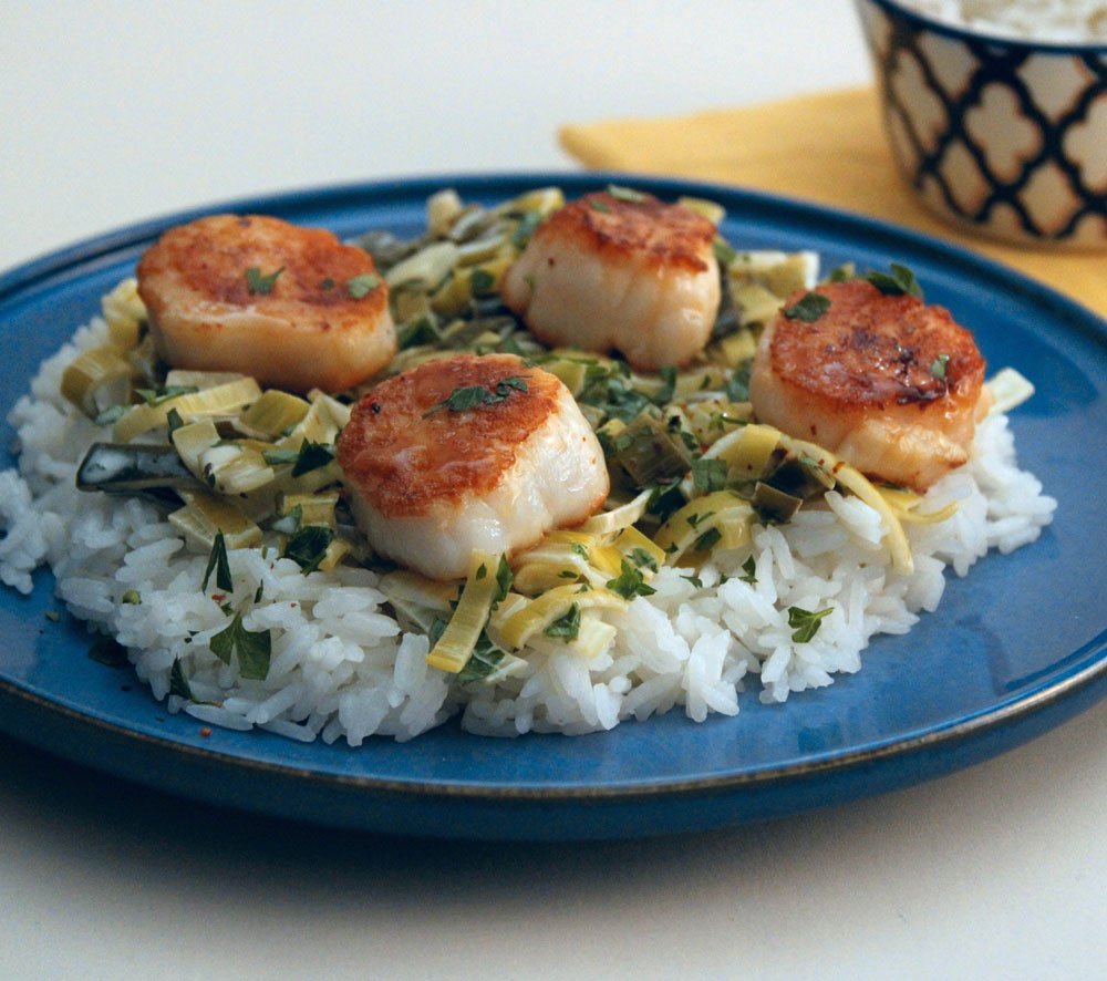Pan-seared scallops served over creamy leeks and fluffy white rice, finished with fresh herbs on a blue plate.
