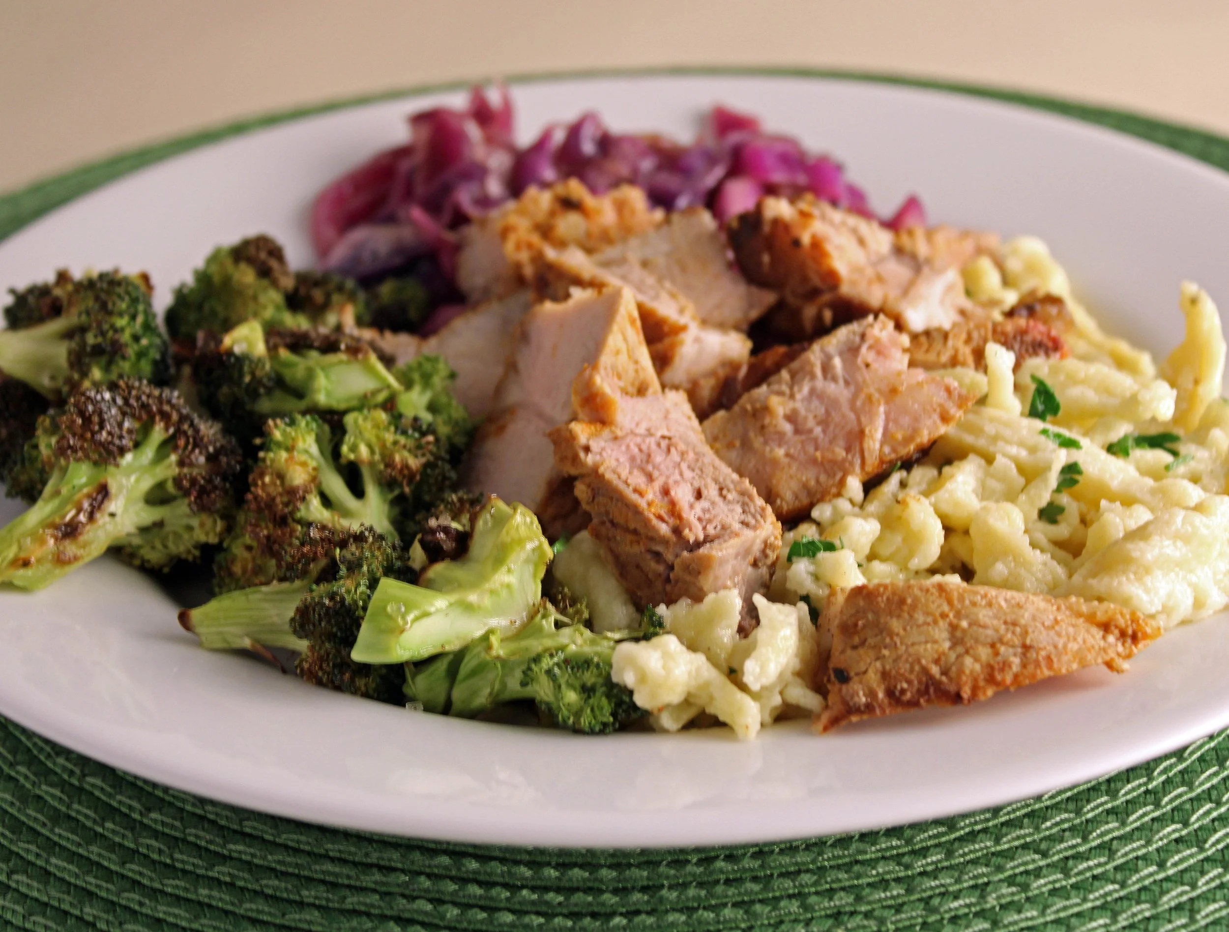 A white oval plate with roasted broccoli, diced roasted turkey, purple coleslaw, macaroni and cheese, and fried fish, resting on a green woven placemat.