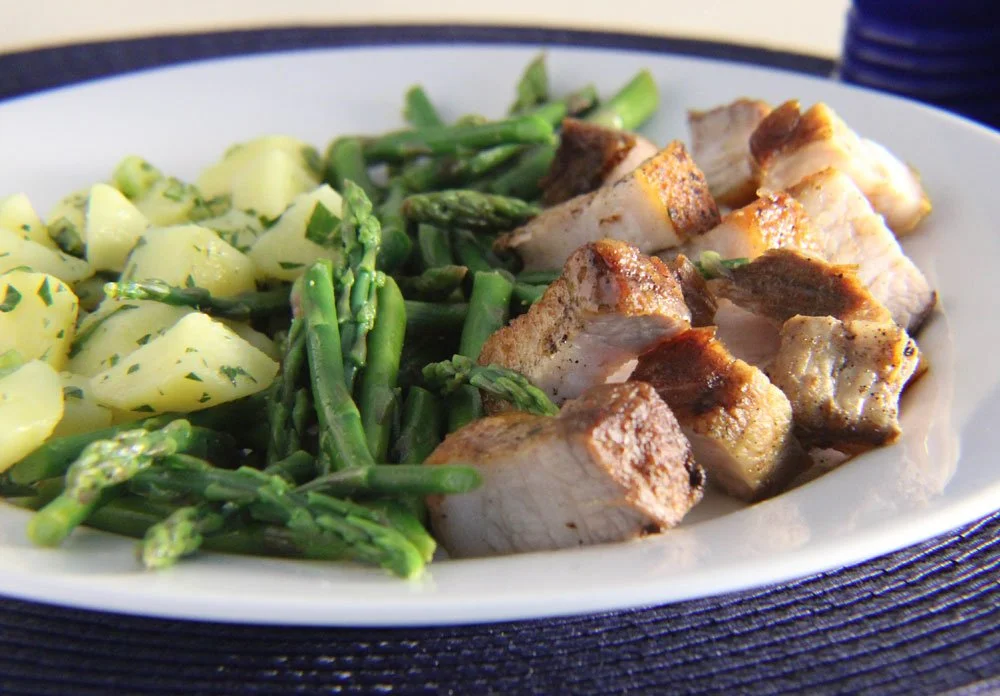 Crispy pork belly with patatas aliñadas and tender asparagus on a white plate.