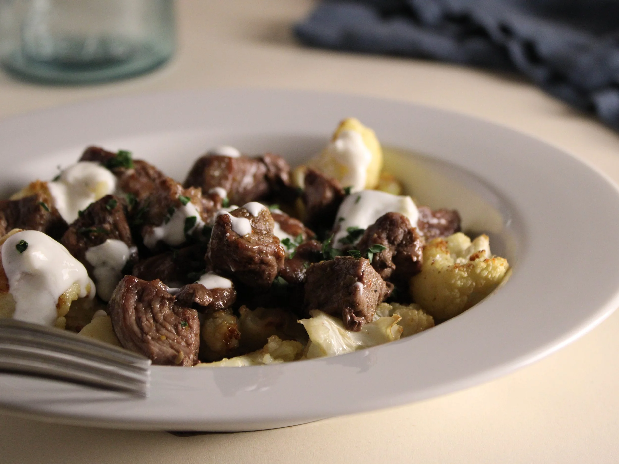 A white bowl with cooked beef chunks, mashed potatoes, and a drizzle of white sauce, sitting on a light-colored surface.