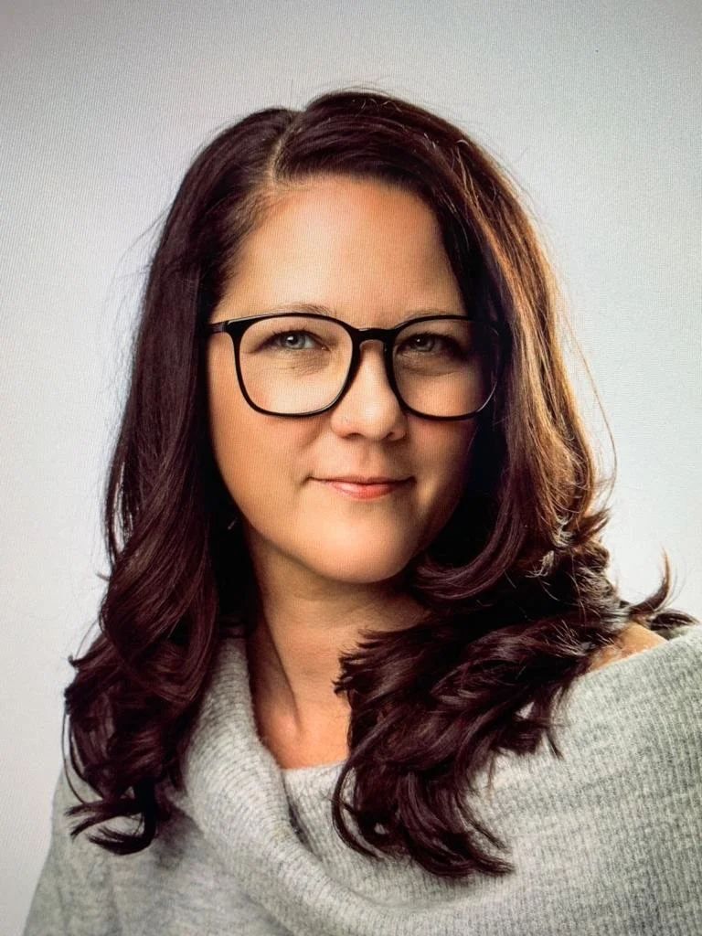 A woman with dark brown, wavy hair and glasses, wearing a gray cowl-neck sweater.