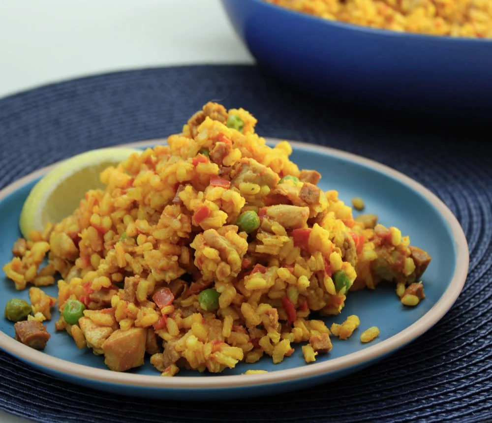 Smoky chicken and chorizo paella served with lemon wedges, peas, and saffron rice on a blue plate with a serving bowl in the background.