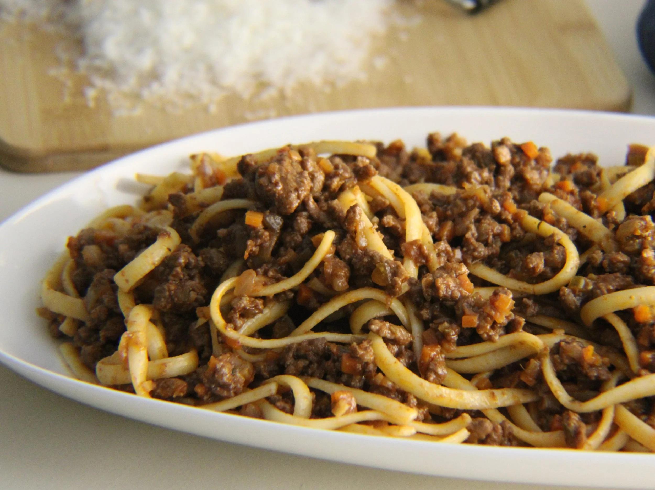 Spaghetti topped with cooked ground meat and diced vegetables in a white bowl, with a container of grated cheese in the background.