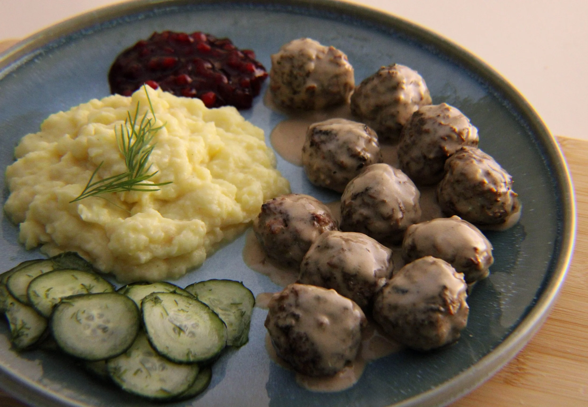 Plate of mashed potatoes garnished with a sprig of dill, sliced cucumbers, and various meatballs with sauce.