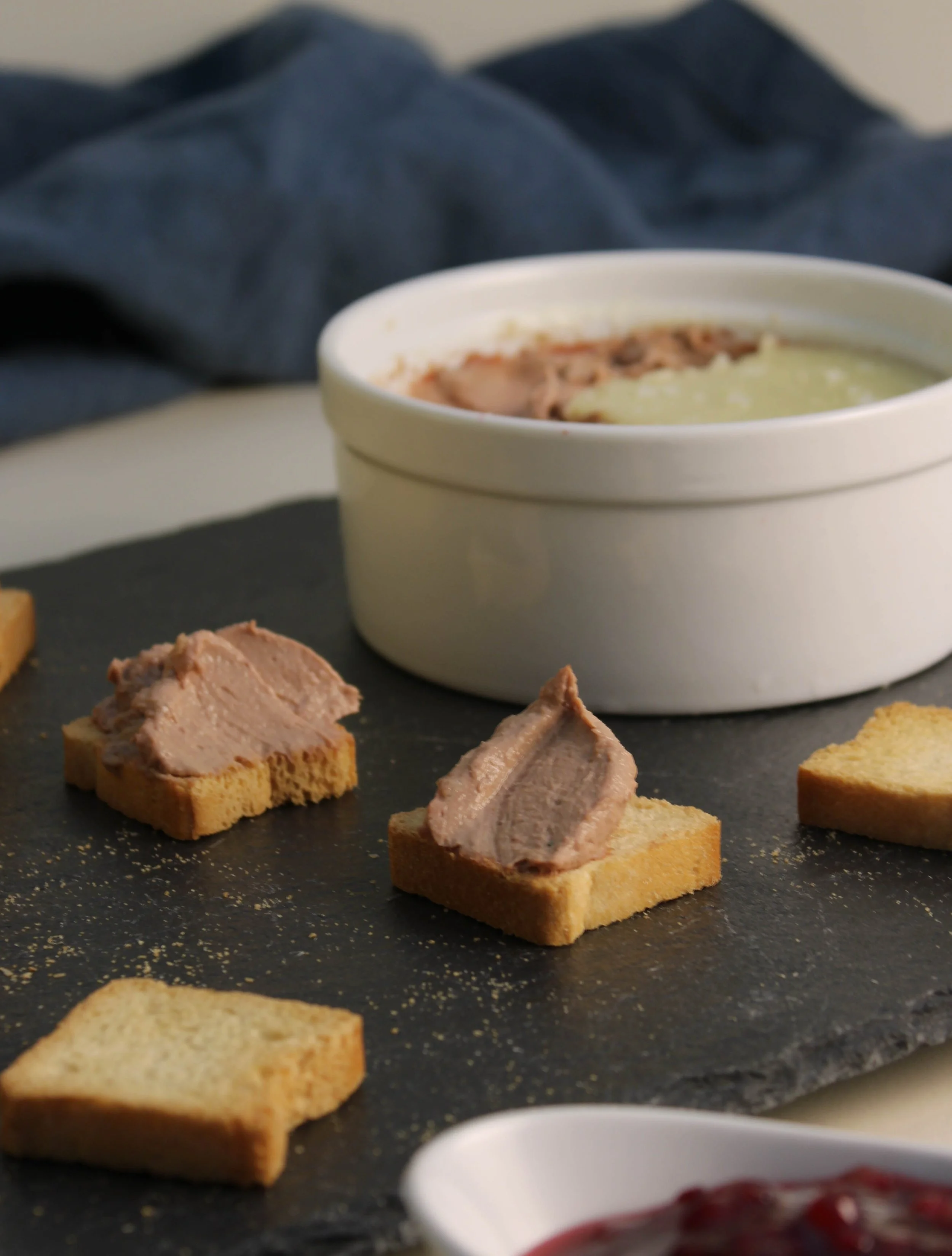 Close-up of thinly sliced pâté on toast, with a black slate serving platter and bowls of spreadable toppings in the background.