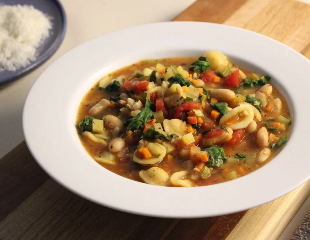 Hearty minestrone soup with white beans, pasta, spinach, tomatoes, carrots and celery in a rich vegetable broth, served in a white bowl.