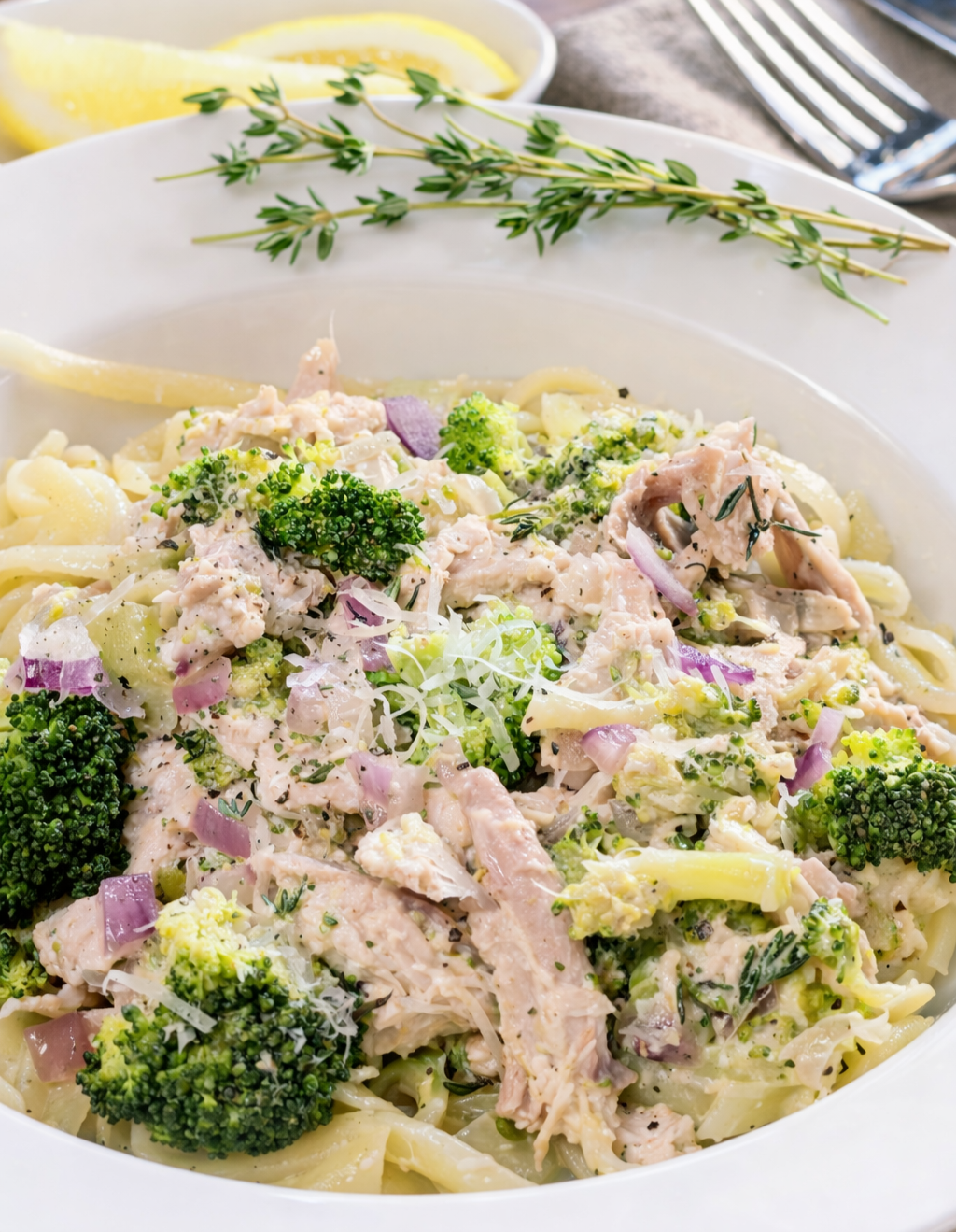 Creamy pasta with broccoli, shredded chicken, red onions, and herbs garnished with thyme sprigs and lemon wedges in the background.