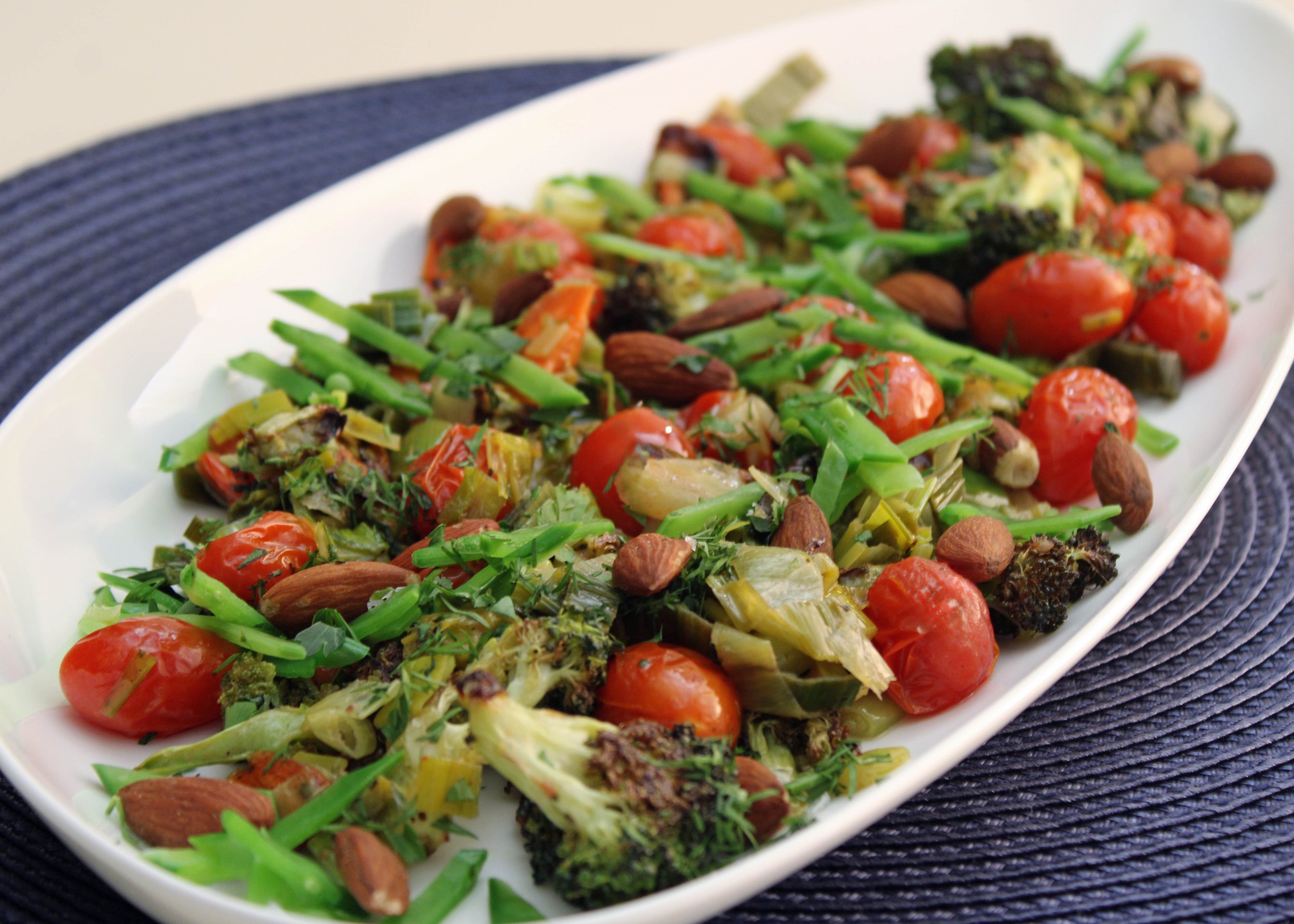 A white oval plate with mixed roasted vegetables including cherry tomatoes, green beans, broccoli, and herbs on a dark woven placemat.