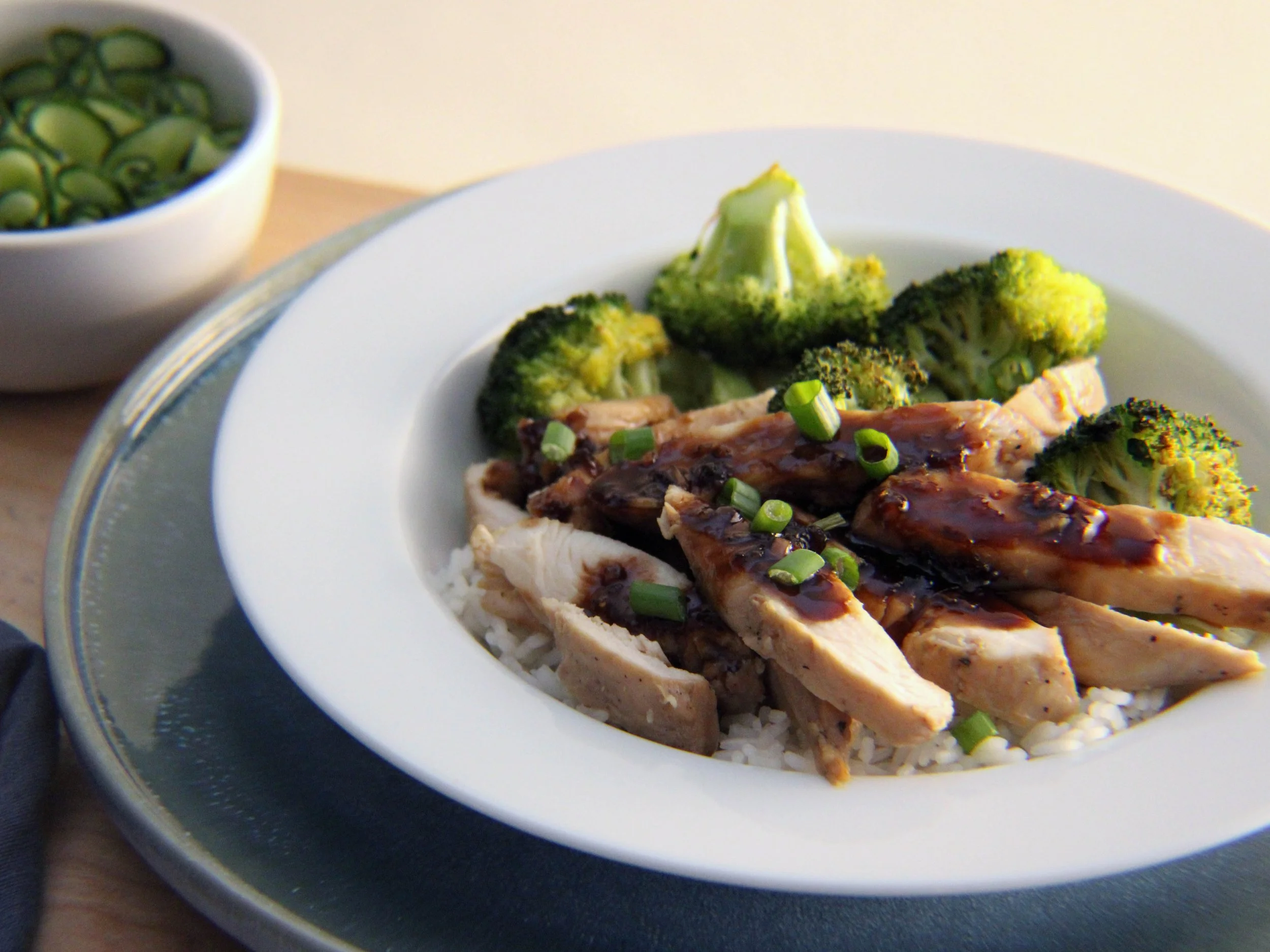 Plate of cooked broccoli with chicken and sauce, with chopped green onions on top, and a bowl of sliced cucumbers nearby.