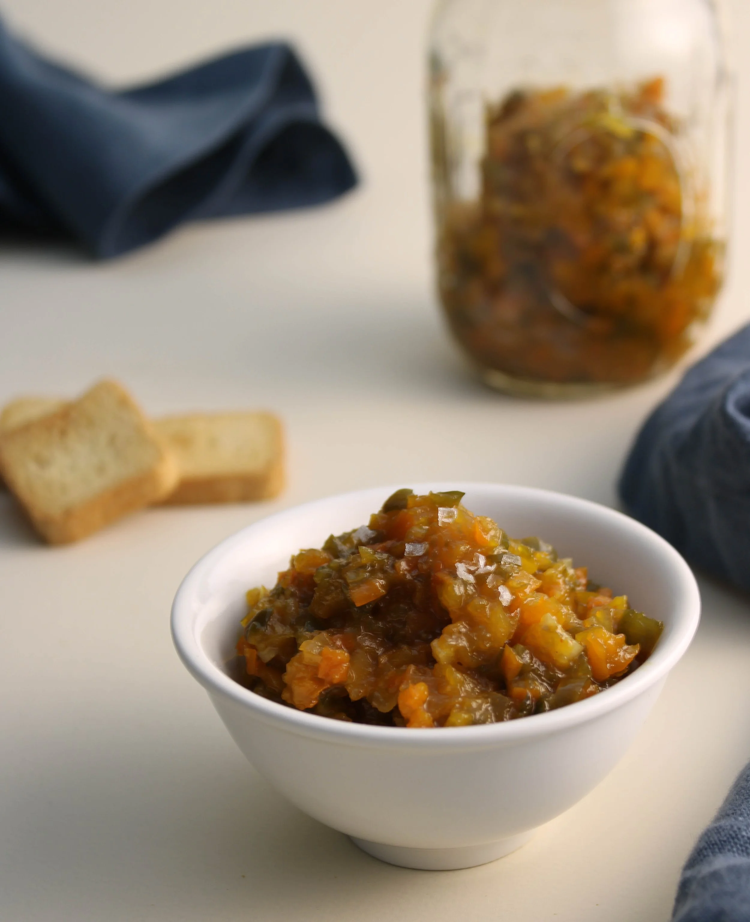 A white bowl filled with cooked vegetable stew, placed on a white surface. In the background, there are two slices of bread on the left, a glass jar of the stew, and a dark blue cloth.