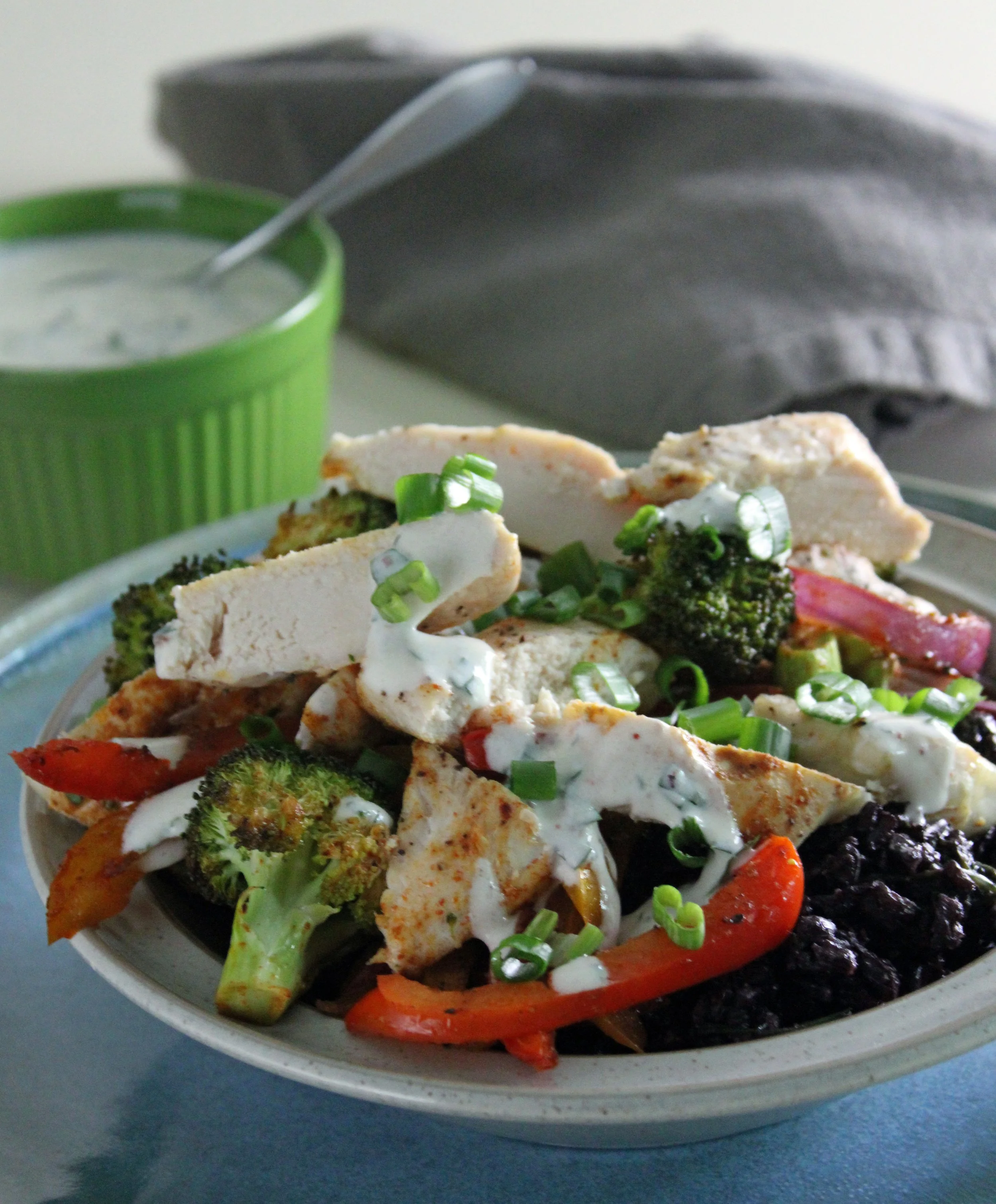 A bowl of black rice chicken bowls topped with lemon chicken, charred peppers, broccoli, spring onions, and a creamy herb yogurt sauce.