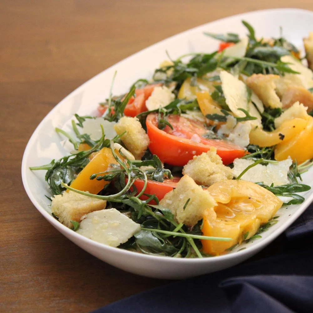 Tomato arugula salad made with heirloom tomatoes, peppery arugula, Parmesan shavings, and crisp croutons, lightly dressed and served on a white platter.