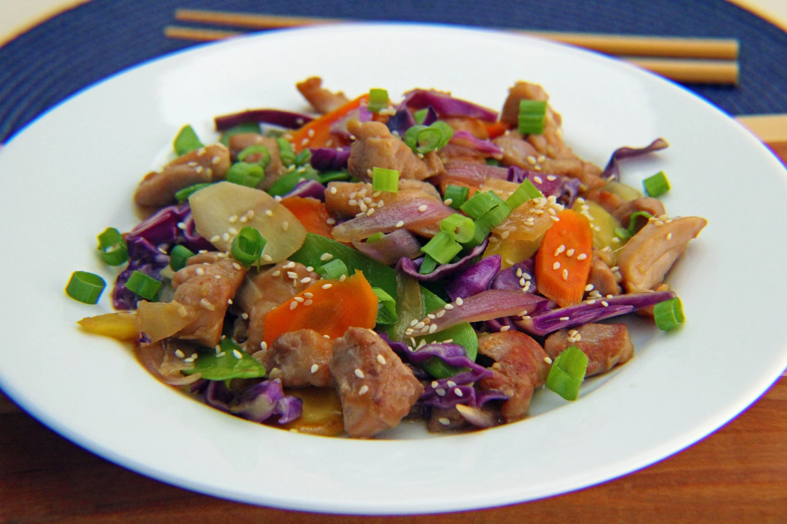A white plate with stir-fried vegetables and diced meat, garnished with chopped green onions and sesame seeds, on a dark placemat and wooden table.
