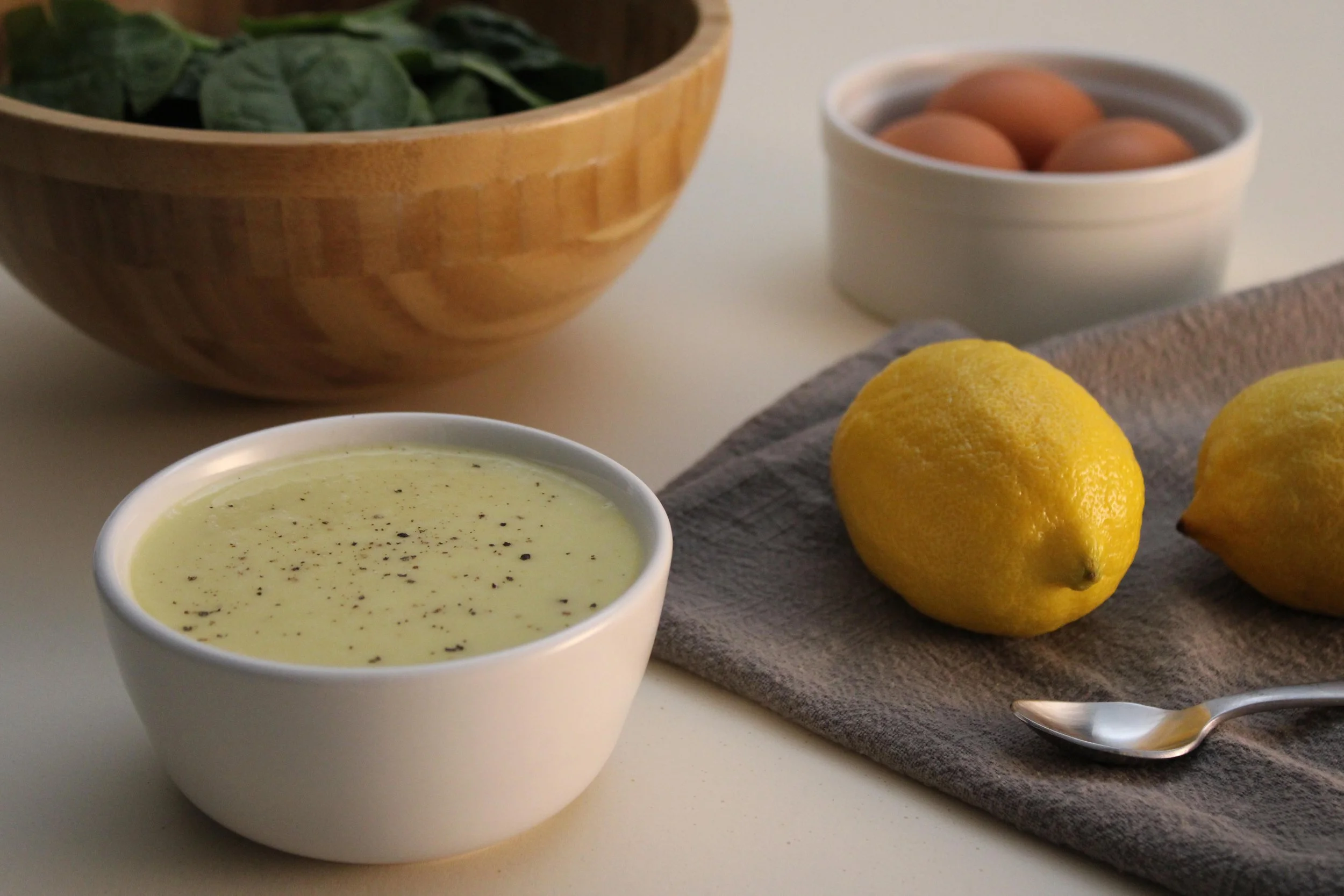 A small white bowl of creamy yellow soup with black pepper on top, two whole yellow lemons on a gray cloth, a bowl of eggs, and a wooden bowl of fresh leafy greens in the background.
