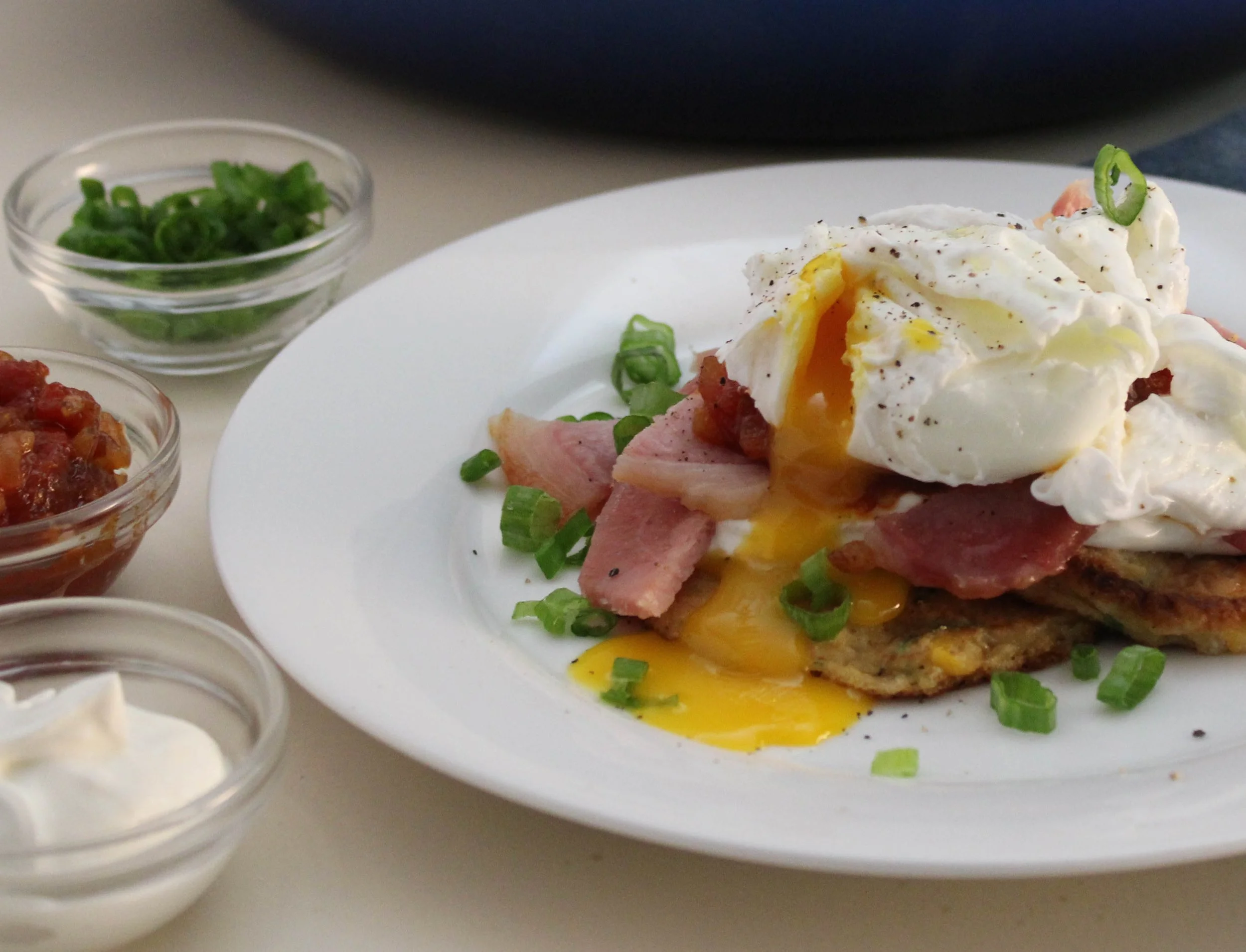 A plate with a poached egg on hash browns, ham, and green onions. Small bowls containing green onions, salsa, and sour cream are nearby.