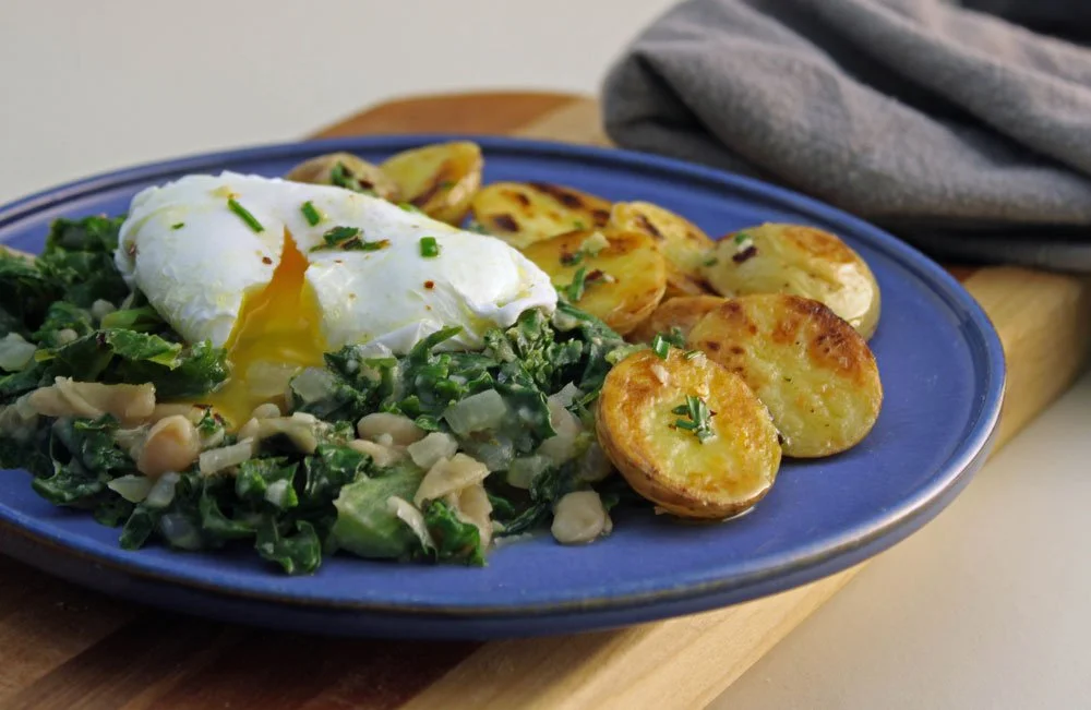 A Tuscan bean stew with cannellini beans, kale, and a creamy poached egg, served alongside crisp golden potatoes for a hearty vegetarian meal.
