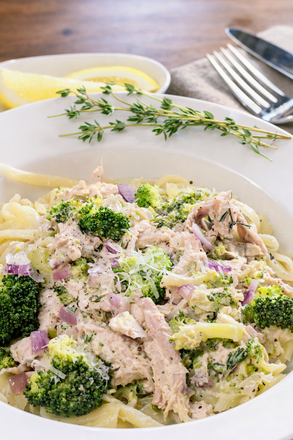 A plate of creamy chicken and broccoli Alfredo pasta garnished with thyme, with lemon wedges and a fork and knife beside it on a gray napkin.
