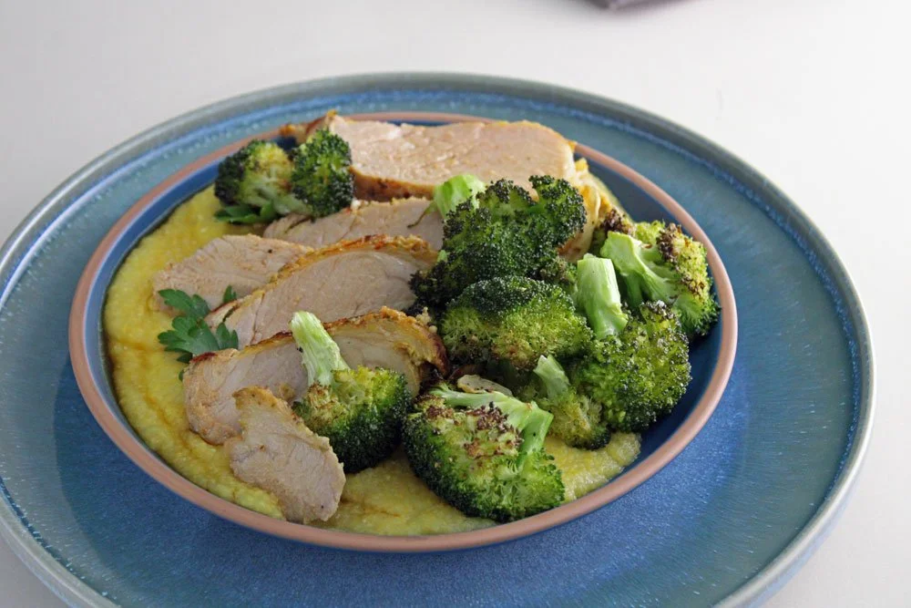 Garlic herb pork tenderloin served over creamy Parmesan polenta with roasted broccoli, pan juices, and fresh parsley in a dark ceramic bowl.