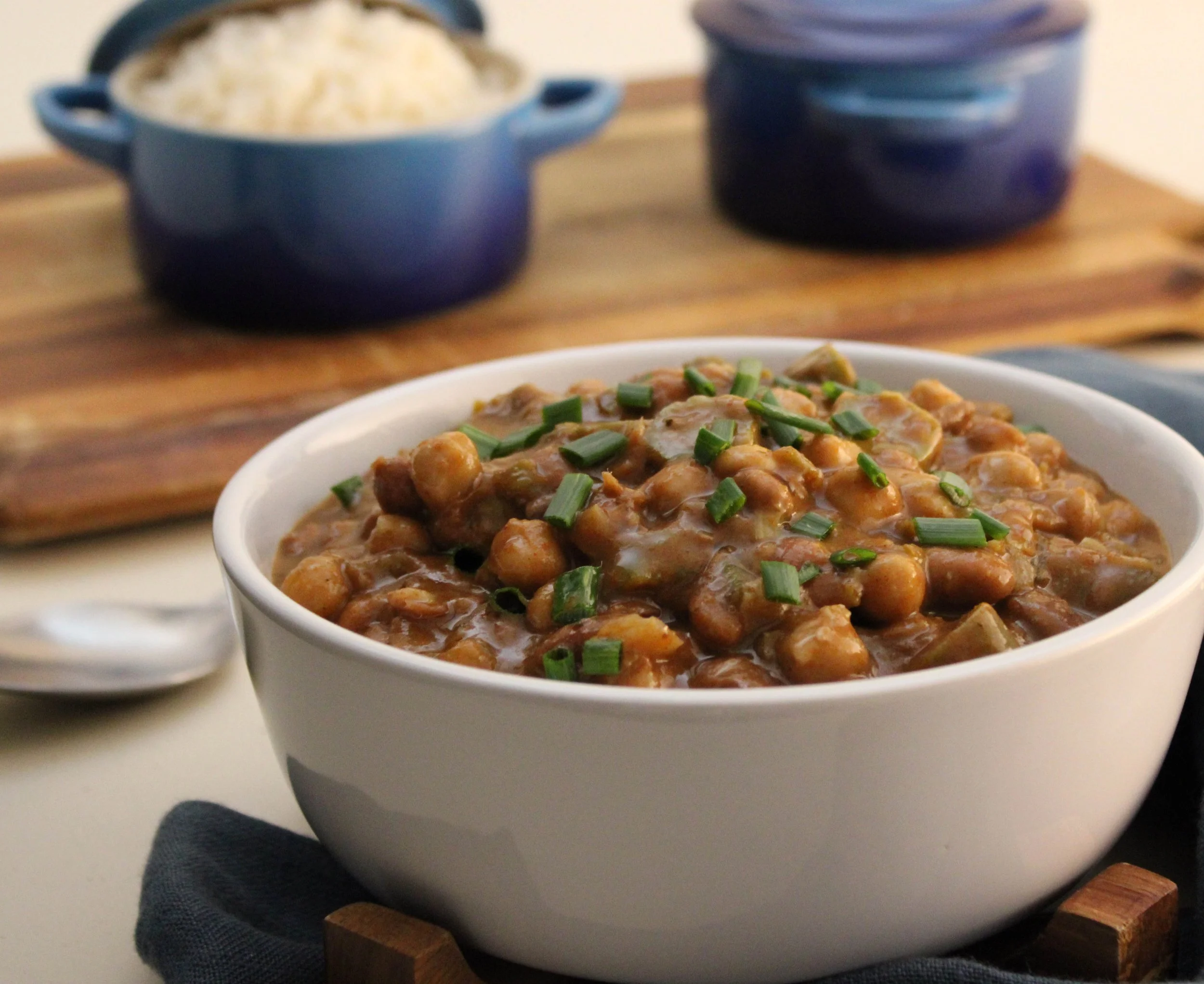 A bowl of chickpea and vegetable stew garnished with chopped green onions in the foreground, with a table of stacked blue bowls and a container of cooked rice in the background.