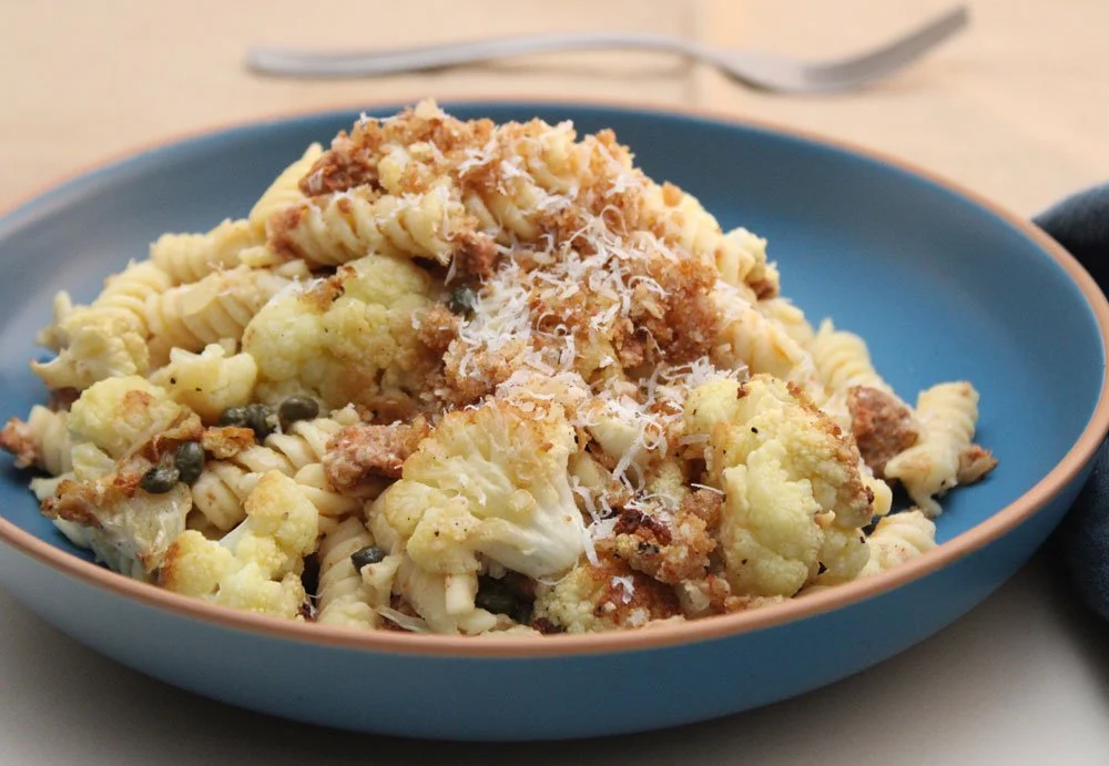 Silken Cauliflower Fusilli with Chorizo and Capers served with roasted cauliflower, whole capers, Parmesan, and crisp golden breadcrumbs in a blue bowl.
