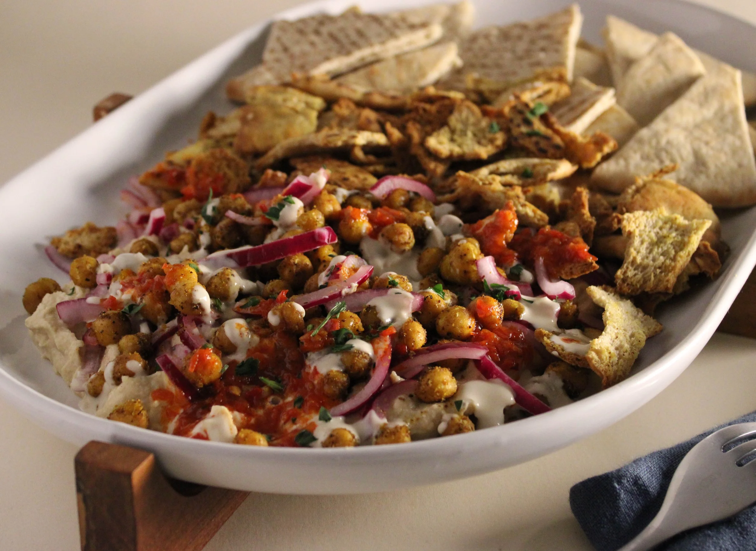 A white oval dish filled with Indian food, including chickpea chaat garnished with chopped onion, cilantro, and a drizzle of yogurt or sauce, with pieces of naan bread around the edge of the dish.