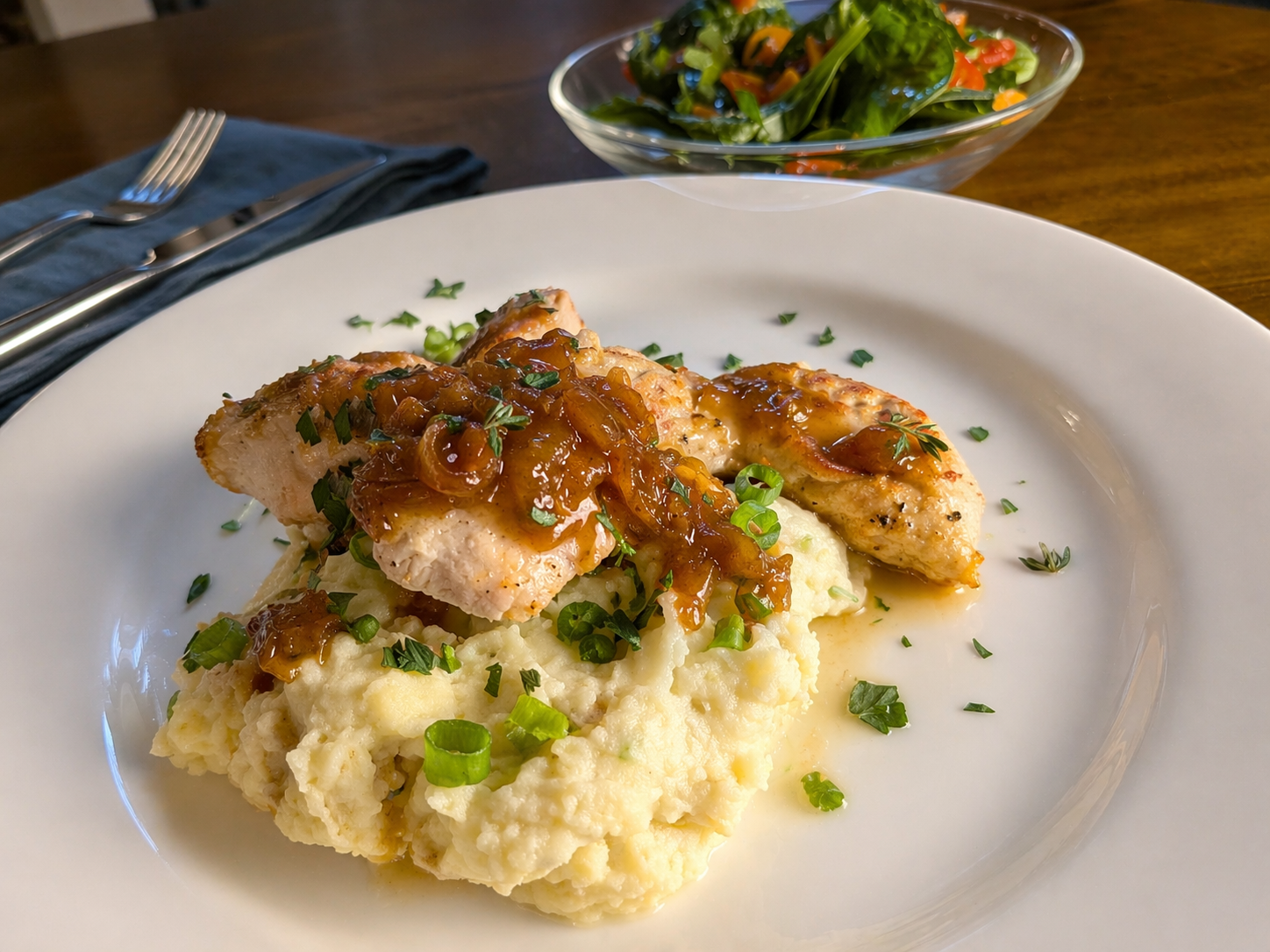 A plate of mashed potatoes topped with grilled chicken and onion gravy, garnished with chopped green onions and parsley, served with a side salad in a glass bowl.