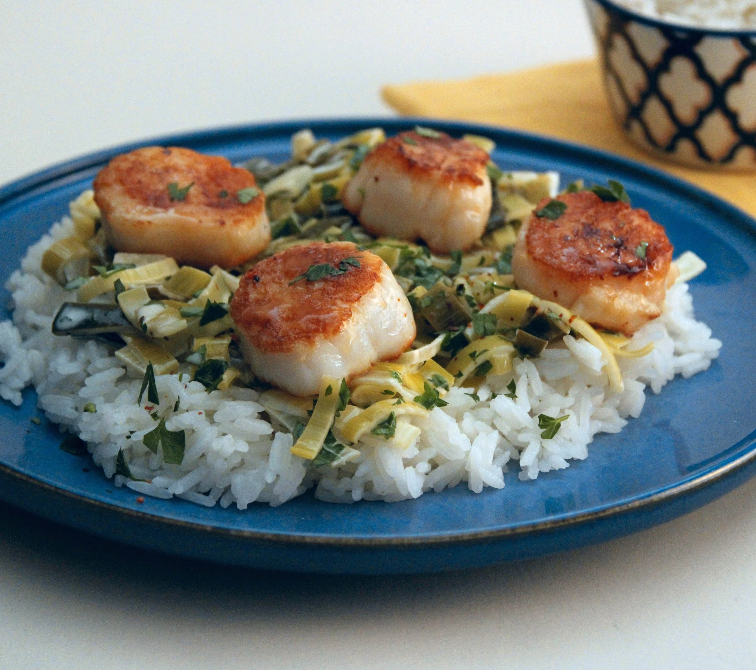 Close-up of seared scallops on a bed of chopped vegetables, garnished with herbs, served on a blue plate.