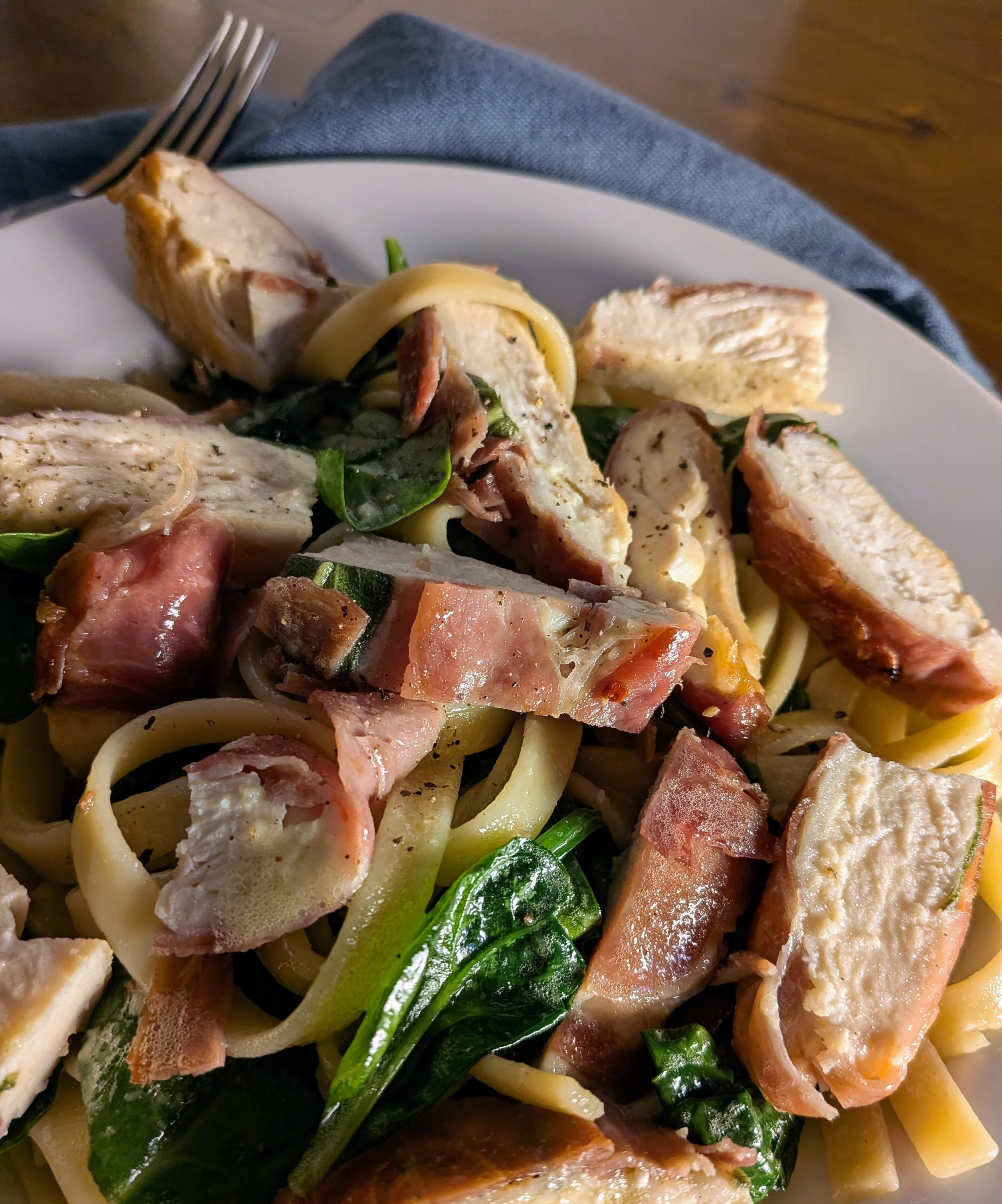 Close-up view of a plate of cooked pasta with chicken, bacon, and vegetables, including zucchini and spinach.