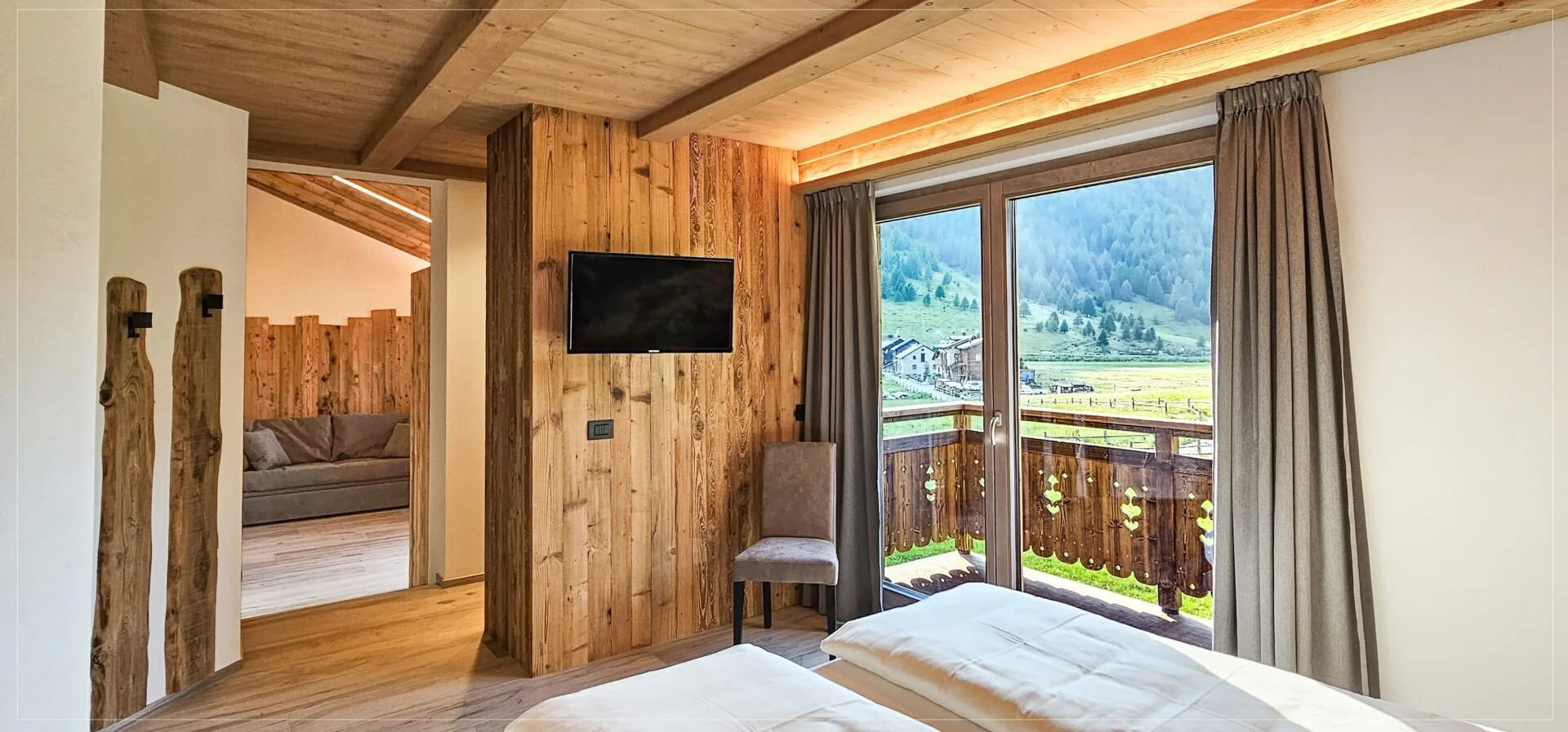 A cozy bedroom with wooden walls, sliding glass door with curtains, and a view of green mountains and houses outside. There is a small chair and a flat-screen TV on the wall.
